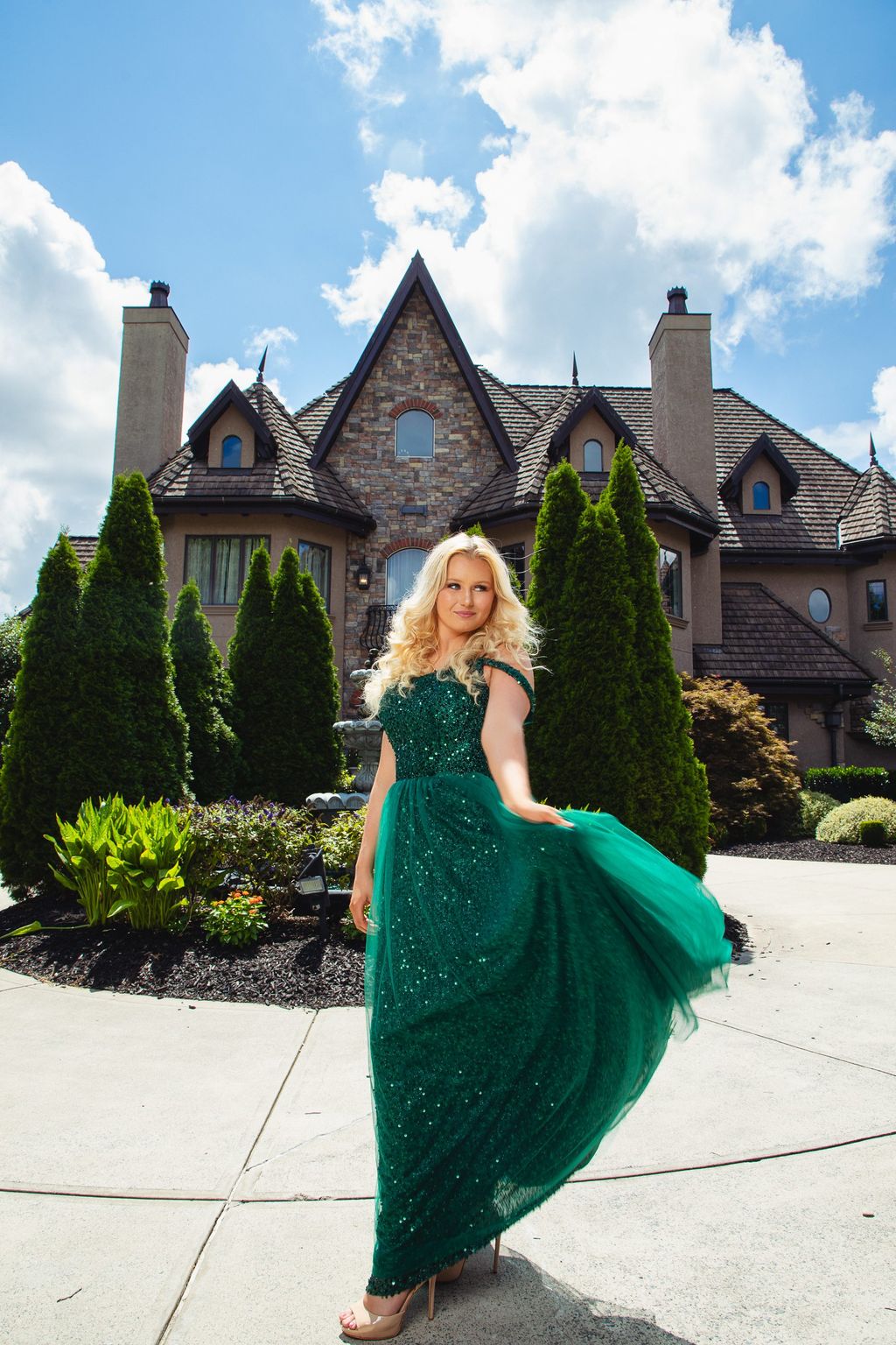 Model in a flowing emerald green gown, arms outstretched, in front of a stone manor under a blue sky.