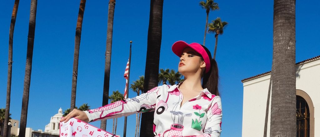 Model in a pink cap and pleated white skirt holding a pink logo tote bag under palm trees.