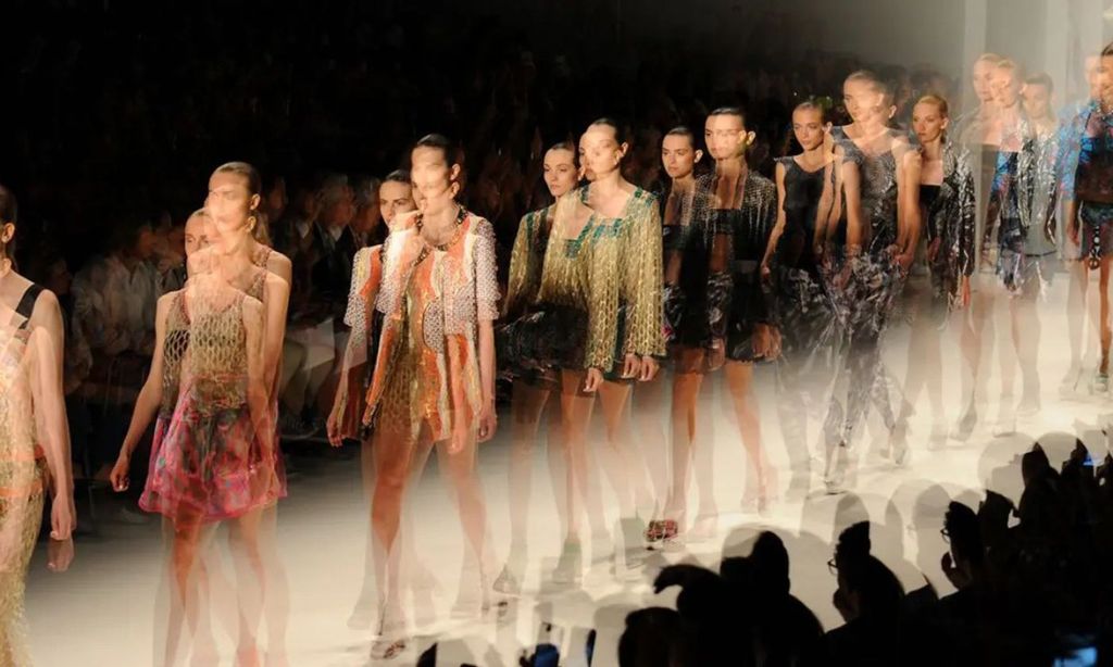 Long-exposure photograph of a runway finale at a fashion week show, models in motion walking past a front row of silhouetted spectators.