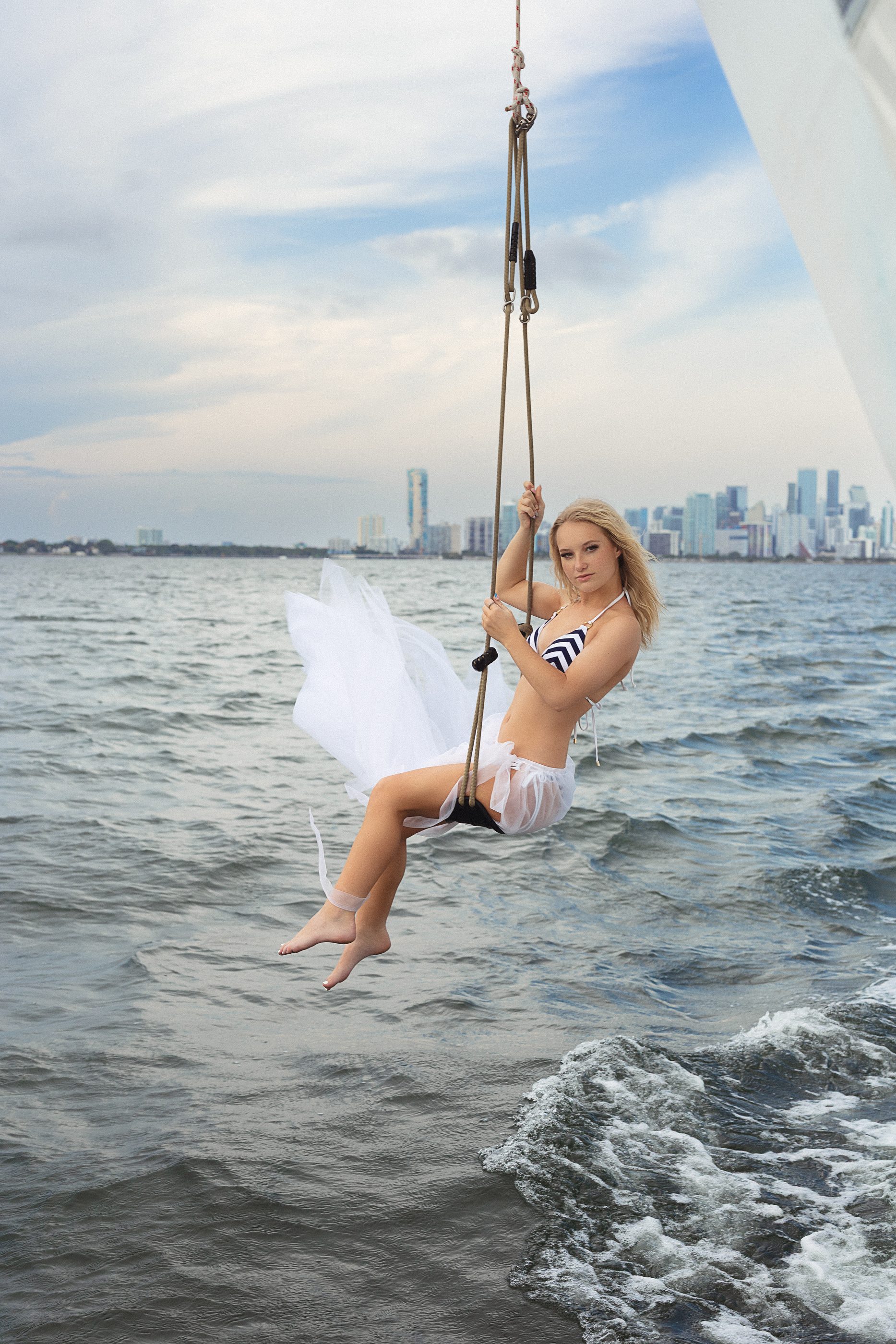 Model in a white tulle skirt on a rope swing over the water, the Miami skyline in the distance.