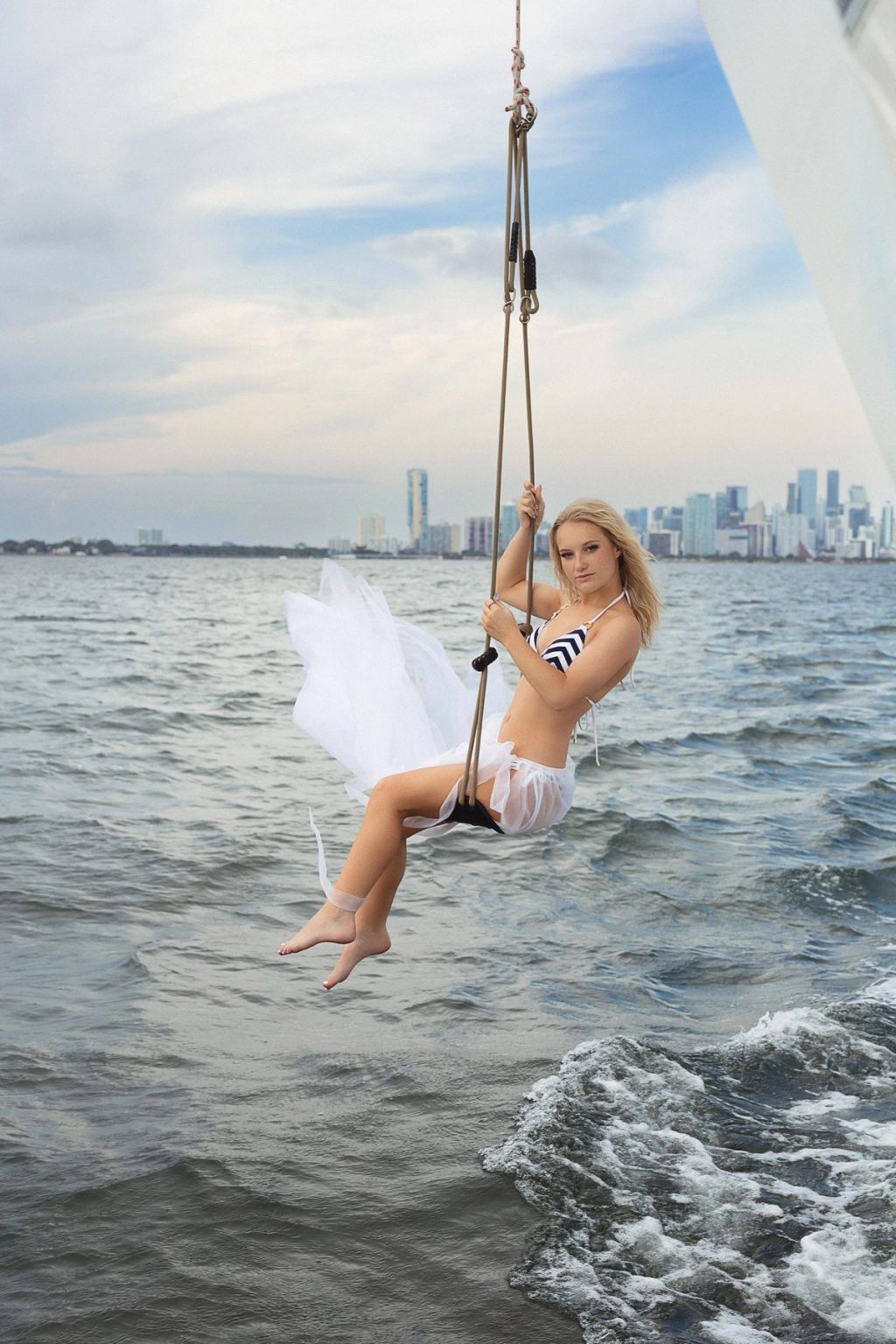 Model in a white tulle skirt on a rope swing over the water, the Miami skyline in the distance.