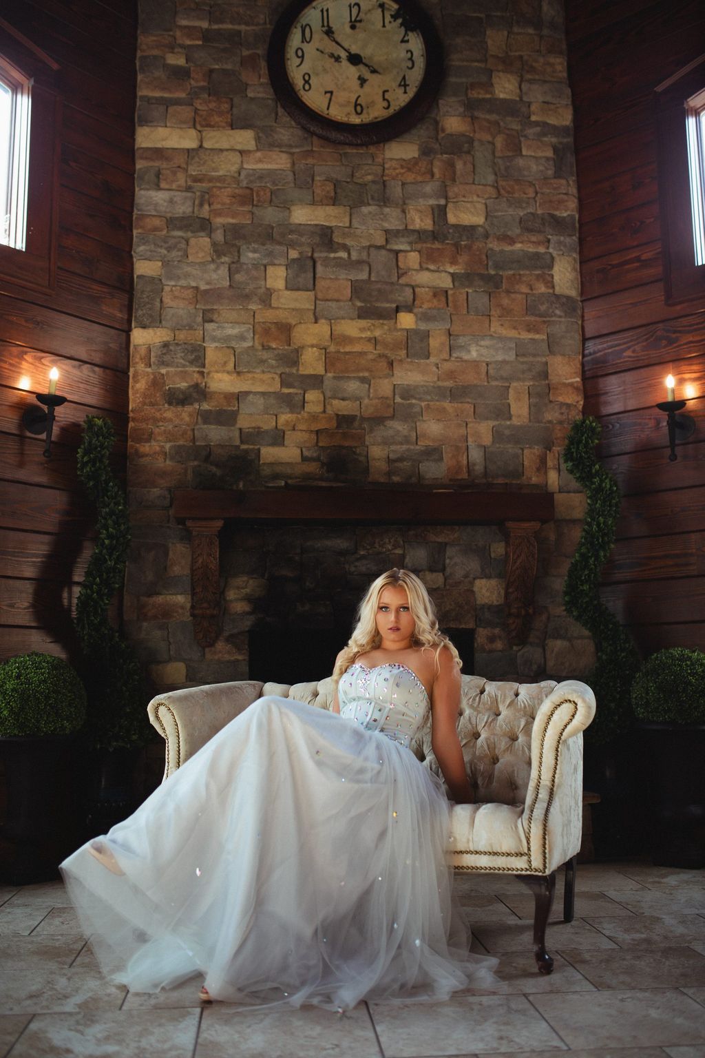 Model in a white tulle gown seated in an armchair beside a grand stone fireplace.