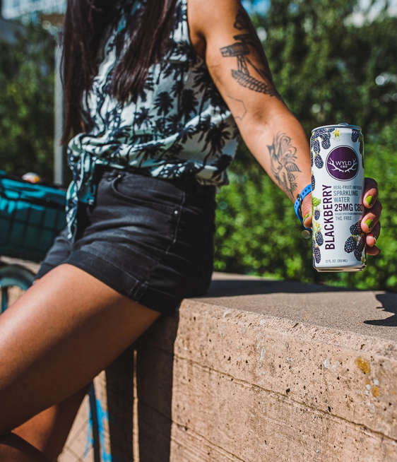 A person standing outdoors in a park holding a can of Wyld Blackberry CBD Sparkling Water.