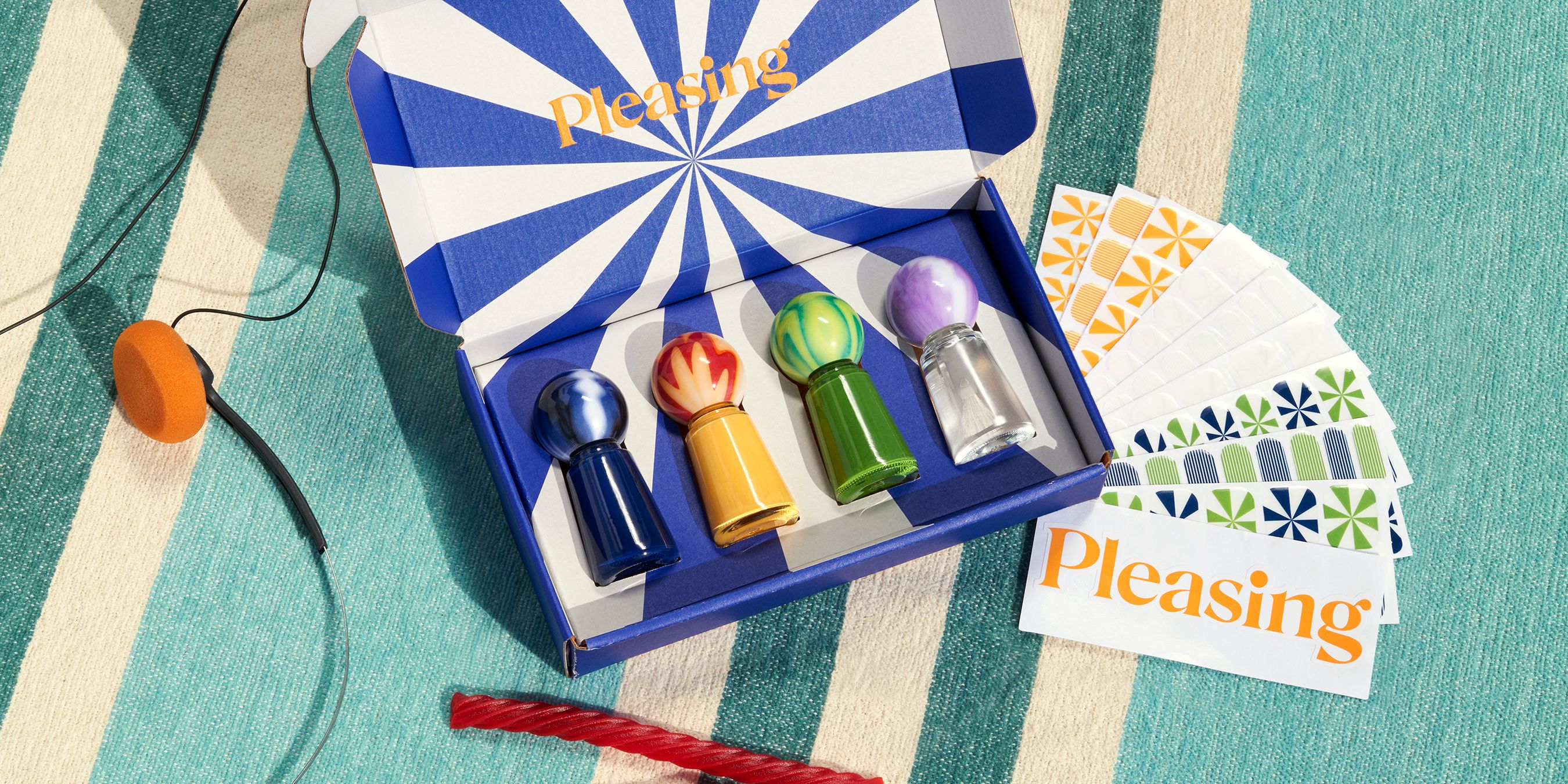 Overhead shot of an open Pleasing gift box with a blue and white sunburst interior holding four colorful nail polish bottles, displayed alongside a fanned set of branded nail art sticker cards on a striped blue and cream surface.