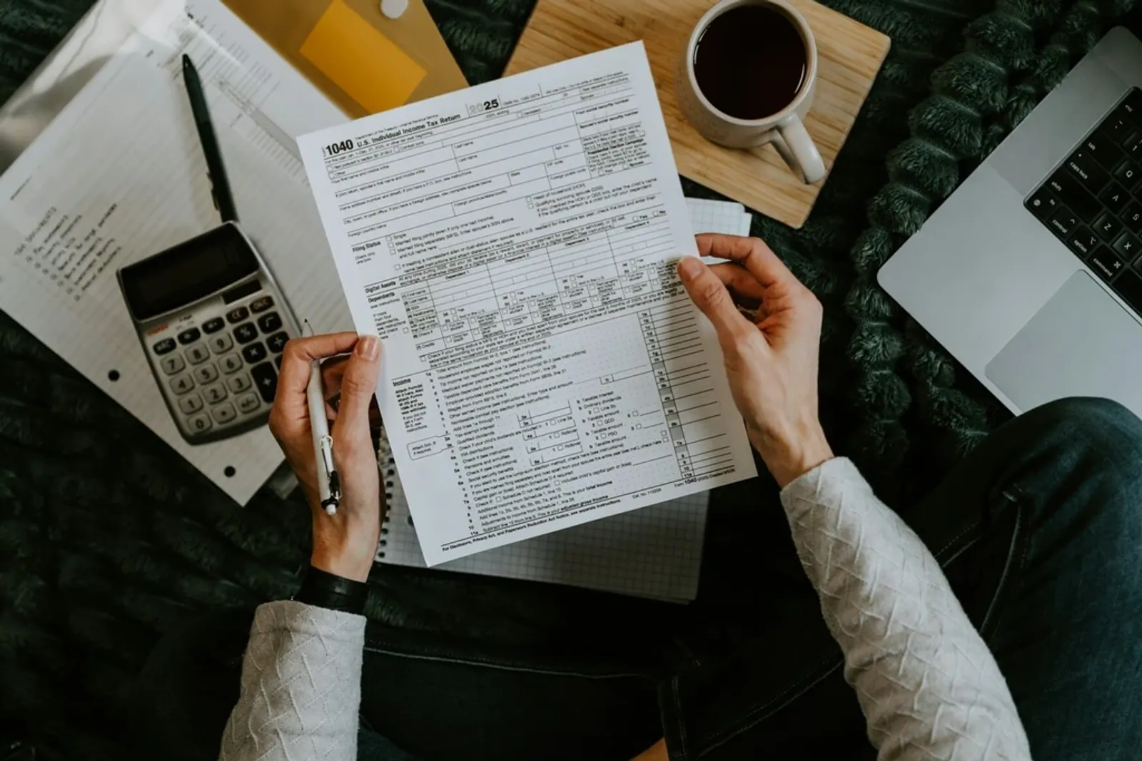 Person filling out a 1040 tax return at a desk with a calculator, coffee, and a laptop.