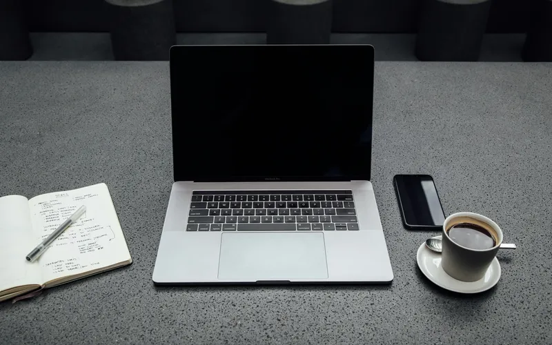 A laptop, open notebook of handwritten notes, phone and cup of coffee on a grey stone desk