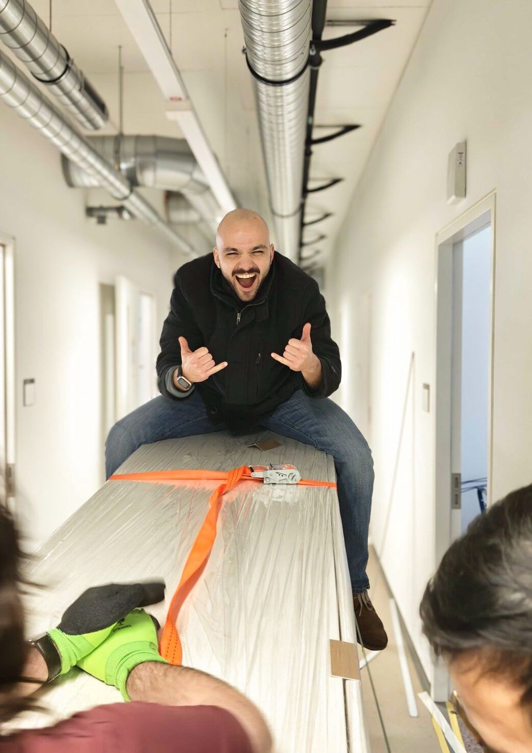 A grinning team member rides a strapped-up package as colleagues move it down a corridor