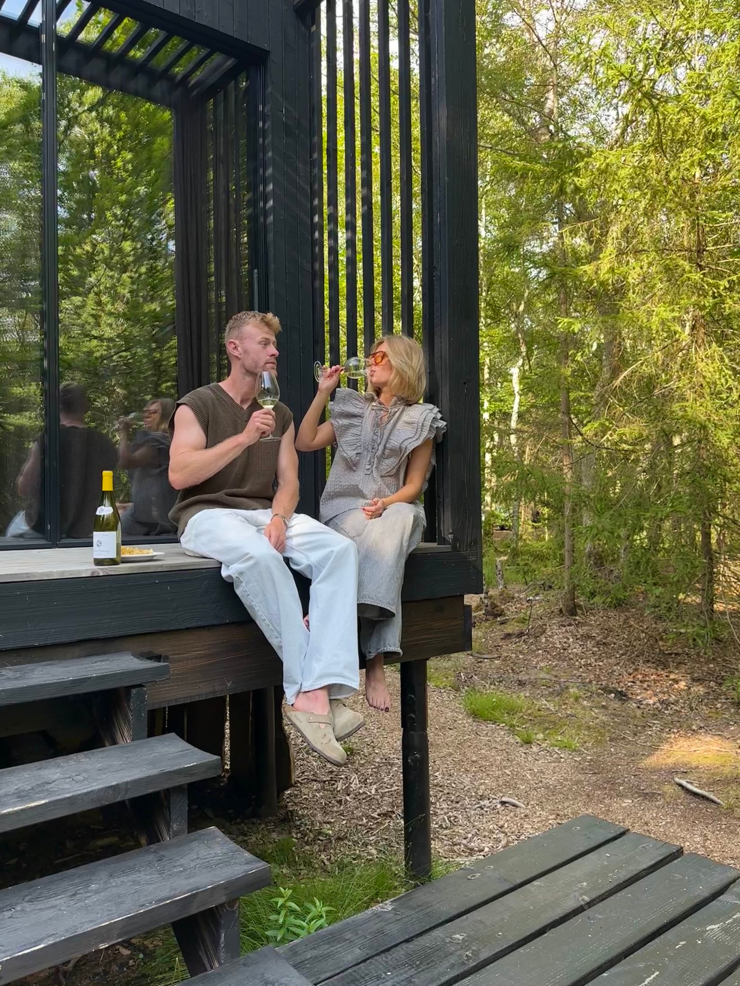 Par drikker vin på terrassen ved en naturovernatning omgivet af skov
