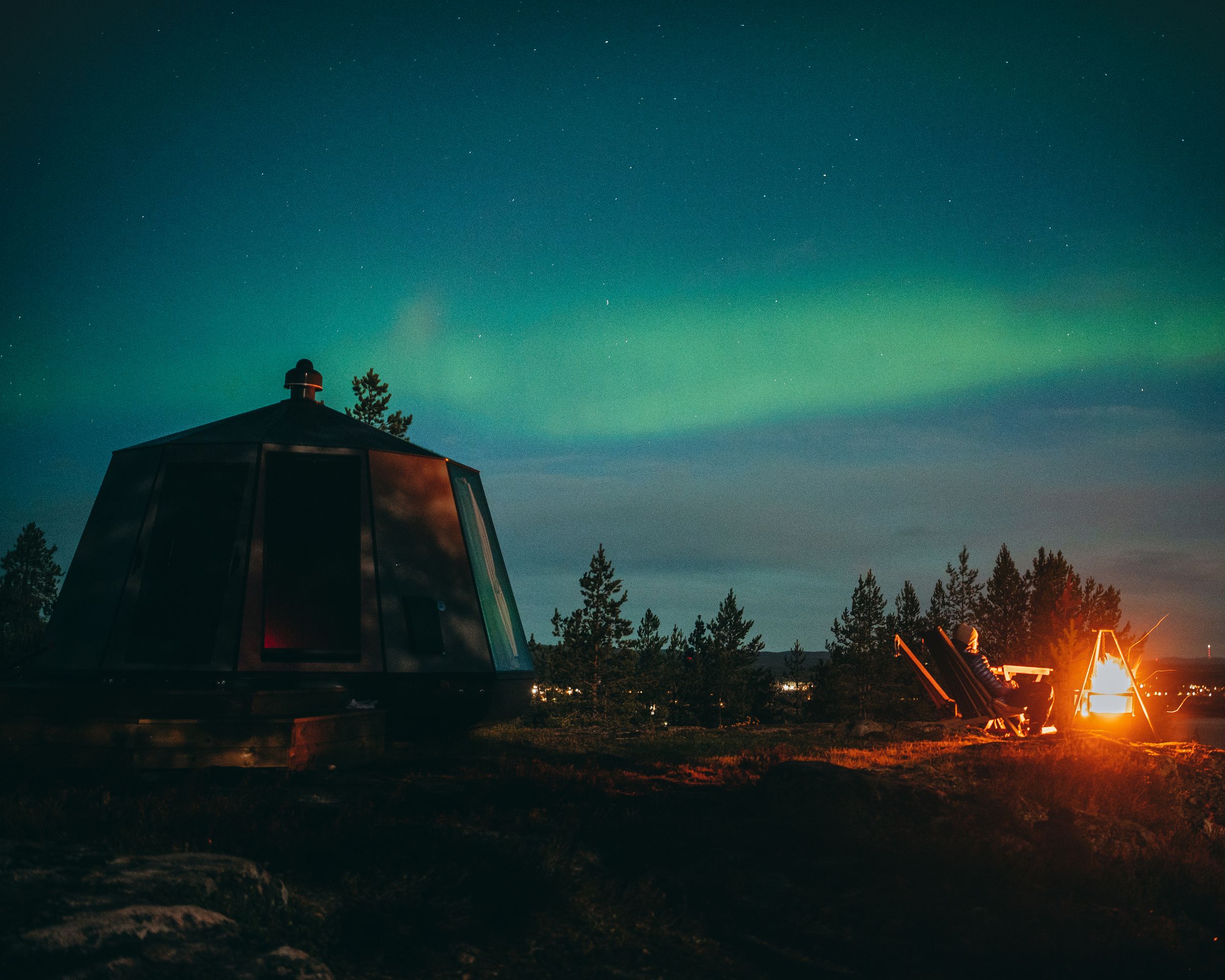 Aurora borealis over a glass dome in Piteå with a man by the fireplace outside