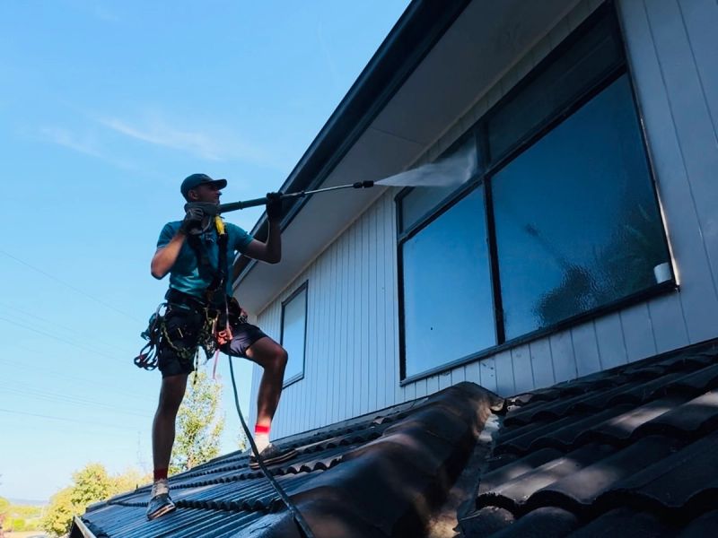 Technician harnessed on a tile roof, washing the eaves and the cladding above the windows from the roof line