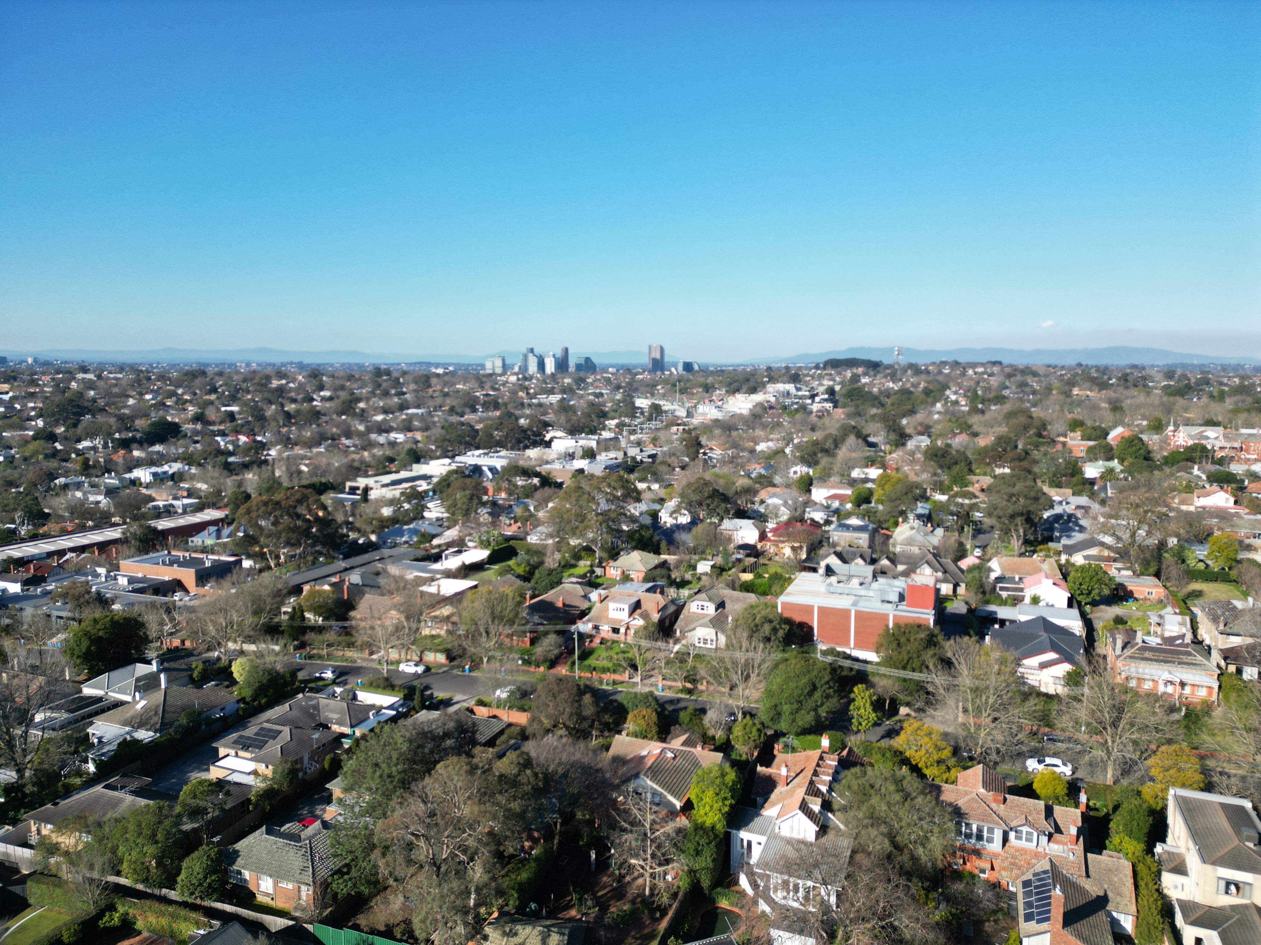 Aerial over Canterbury rooftops packed between mature trees, city skyline on the horizon