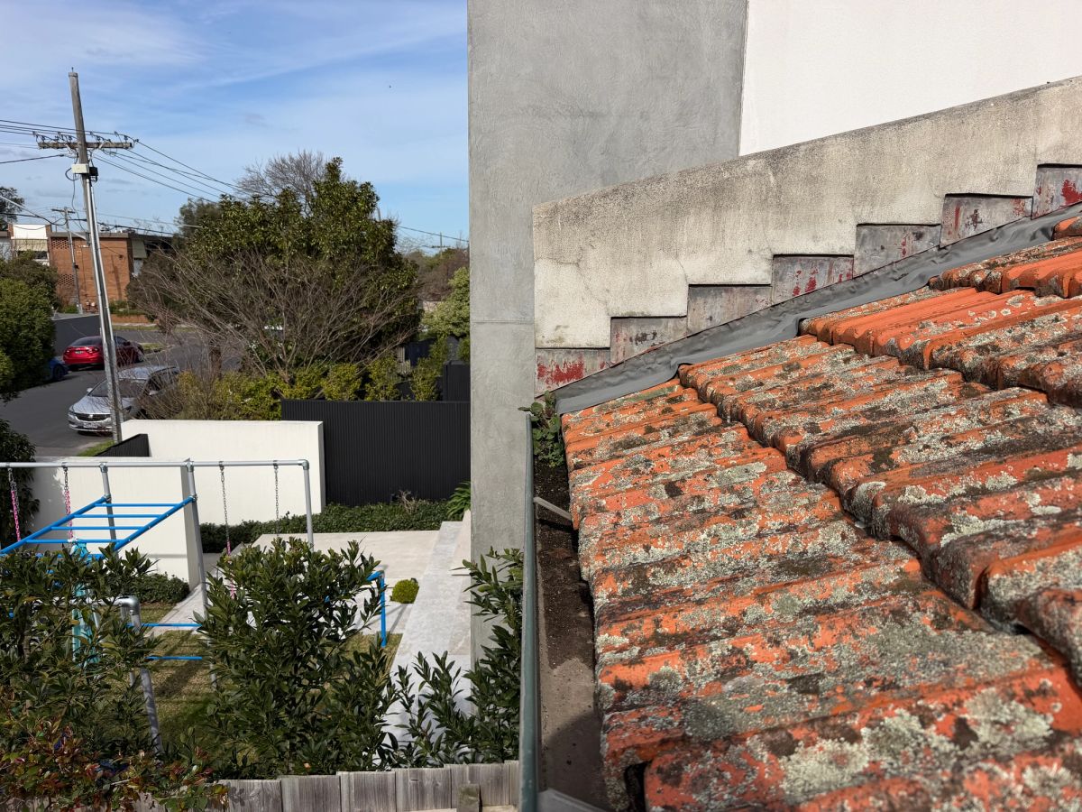 Gutter run against a rendered wall on Hoddle Street, Elsternwick, filled with compacted debris beside a lichen-covered terracotta roof