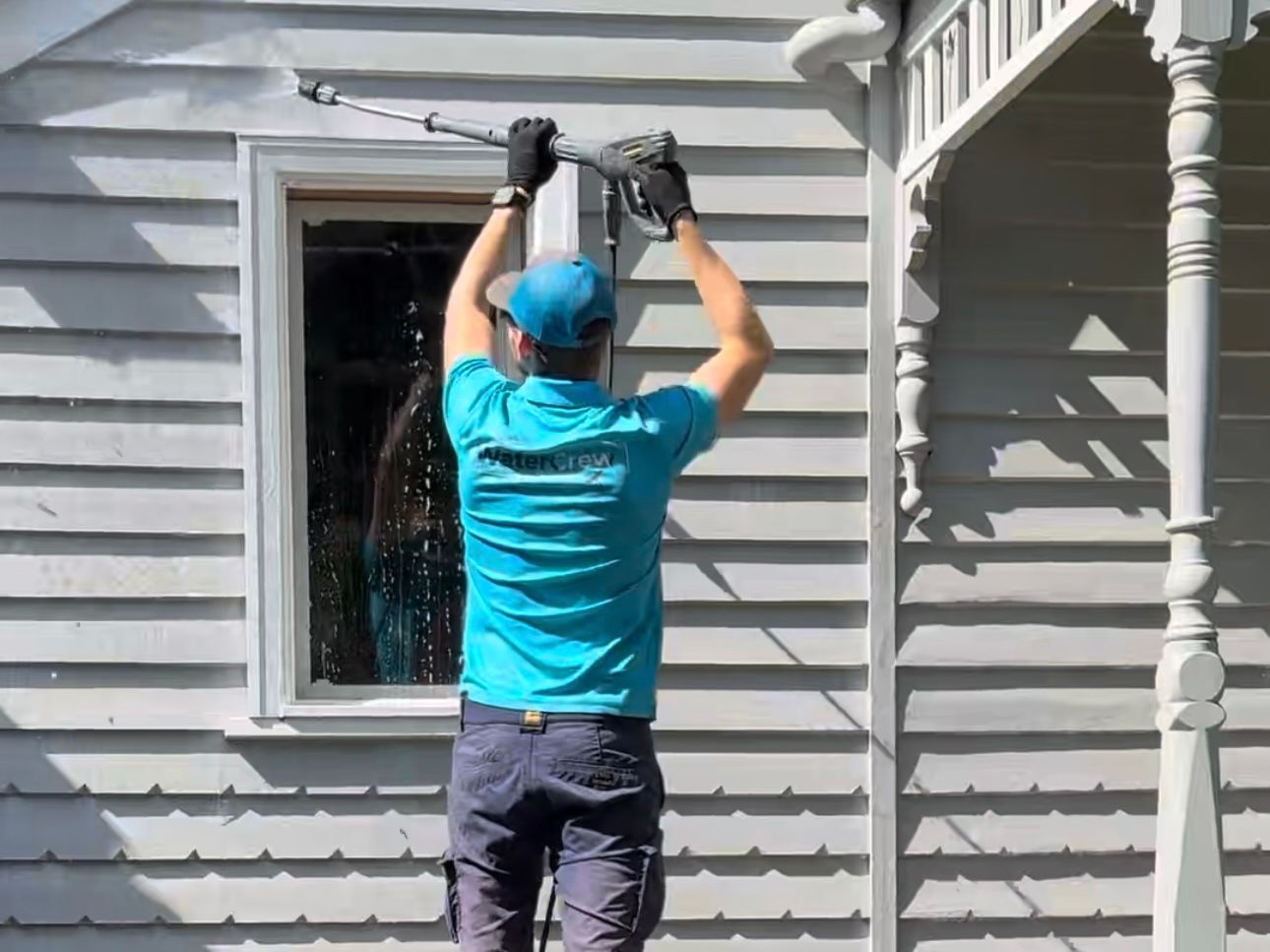 WaterCrew technician washing grey weatherboard cladding and a window surround beside an ornate verandah post