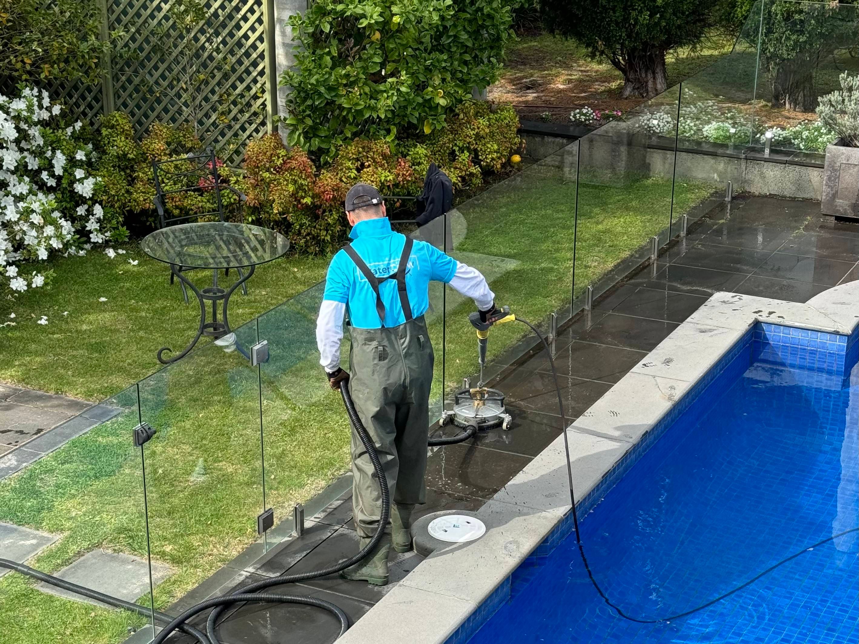 Technician in waders cleaning a wet bluestone pool surround behind a glass pool fence