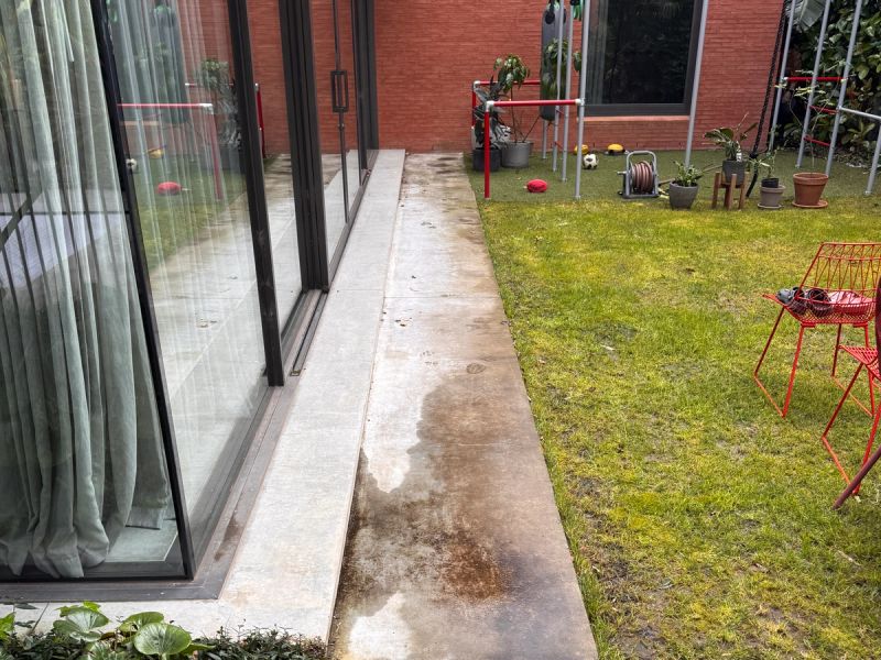 Concrete path beside a glass wall, dark staining along the outer edge against the lawn