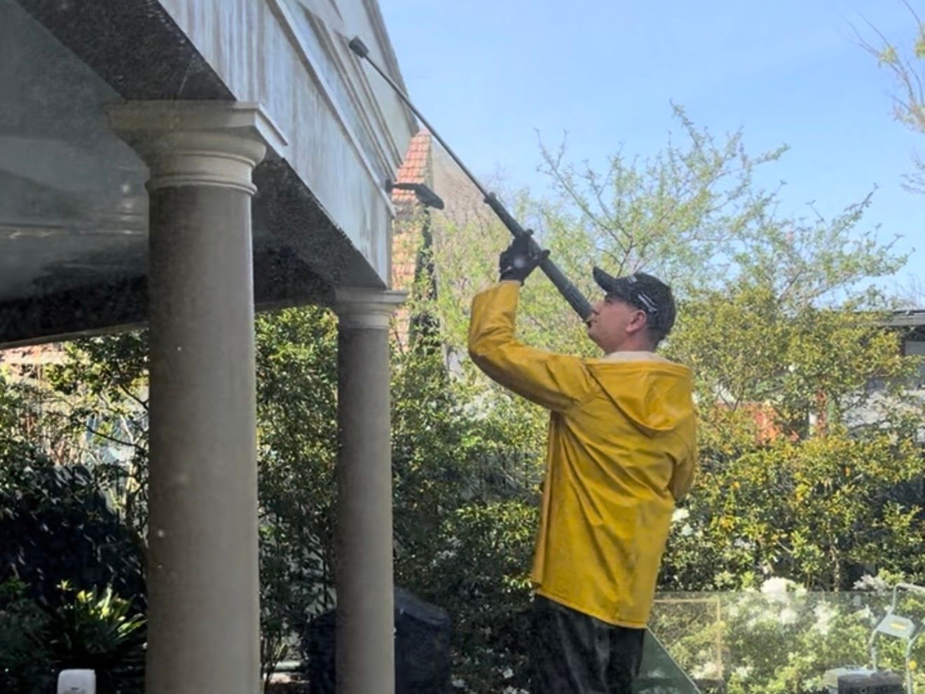 Technician in wet-weather gear washing the eaves above a rendered portico, water running down the columns