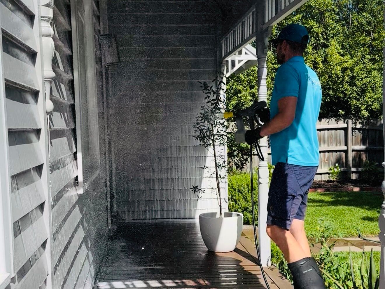 Technician in a WaterCrew polo rinsing dark weatherboard along a side verandah, water sheeting down the boards