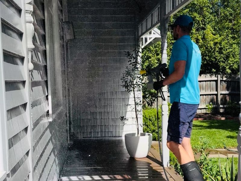 Technician in a WaterCrew polo rinsing dark weatherboard along a side verandah, water sheeting down the boards