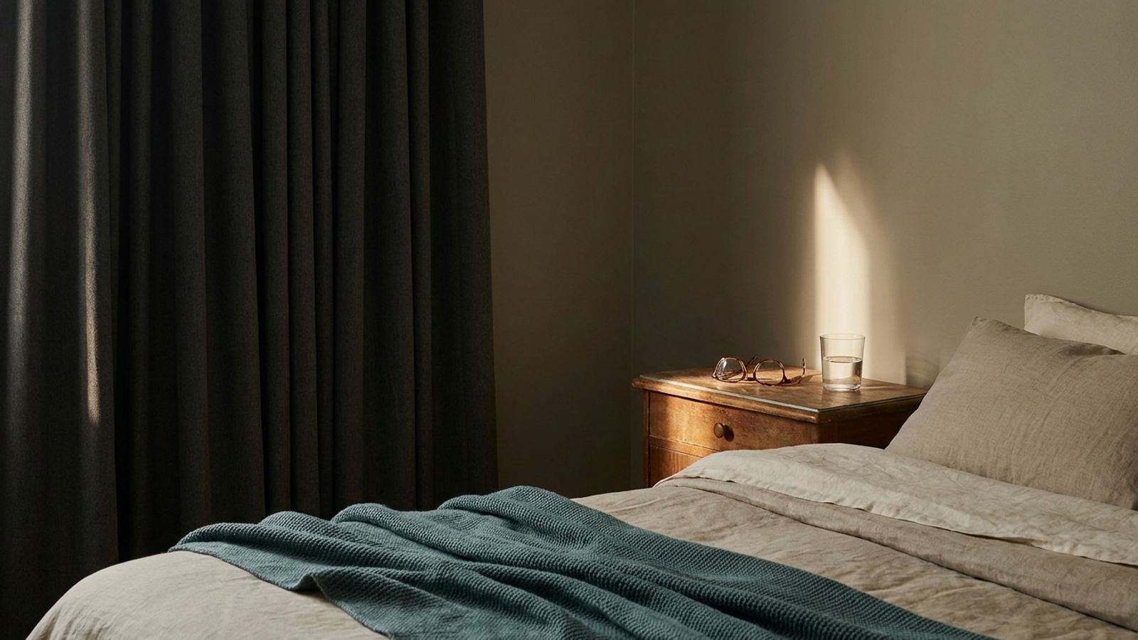 A dim bedroom with dark curtains, glasses and a glass of water on the nightstand, and a teal throw