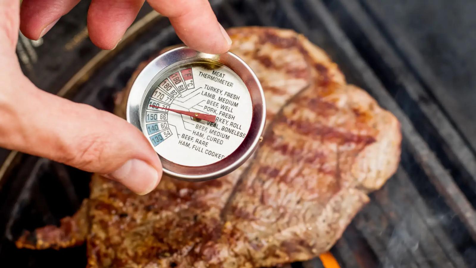Hand misst Kerntemperatur eines Steaks auf dem Grill mit analogem Fleischthermometer