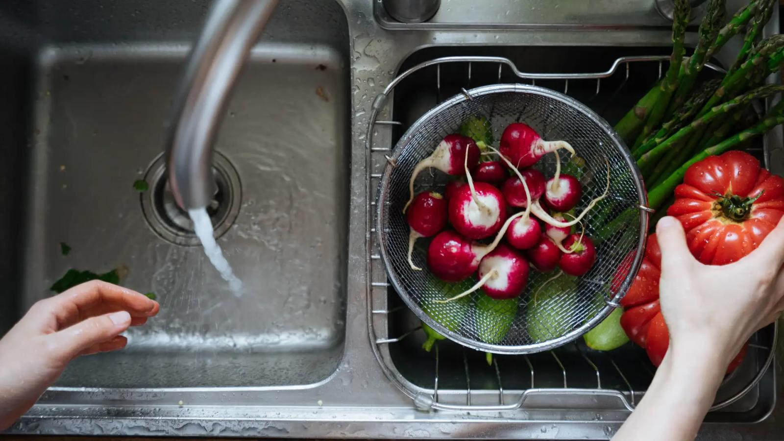 Frisches Gemüse wie Radieschen, Spargel und Tomate wird in einem Sieb unter fließendem Wasser in der Edelstahl Küchenspüle gewaschen