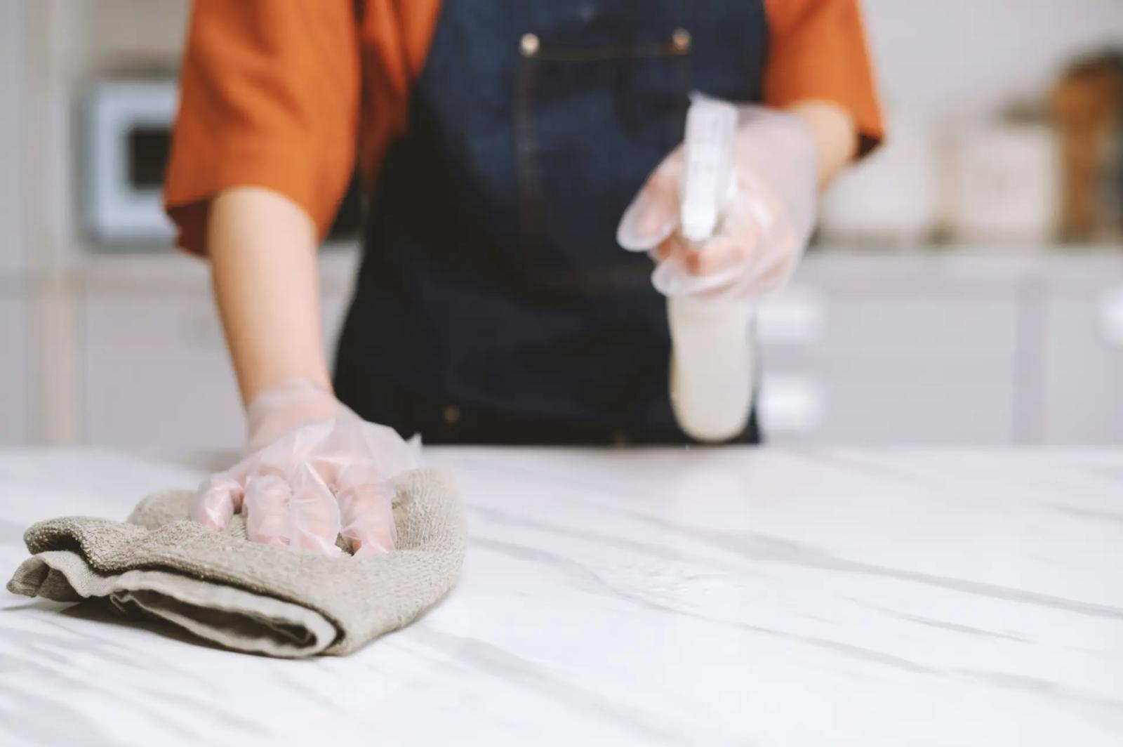 Person reinigt eine weiße Marmor-Arbeitsplatte mit Mikrofasertuch und Sprühreiniger, trägt dabei durchsichtige Handschuhe, Küchenarbeitsplatte richtig reinigen ohne Rückstände