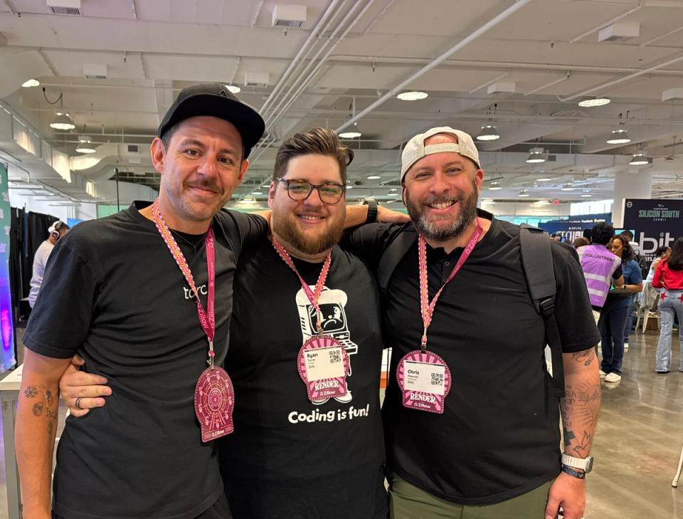 Alt text:  Three men standing closely together indoors at a conference or event, smiling at the camera. The man on the left wears a black cap and black t-shirt with a pink lanyard and badge. The man in the middle, wearing glasses and a black t-shirt that says "Coding is fun!" with a computer graphic, has his arms around the other two. The man on the right wears a white backward cap, black shirt, and backpack, also with a pink lanyard and badge. The background shows other attendees and booths in a large, well-lit room.