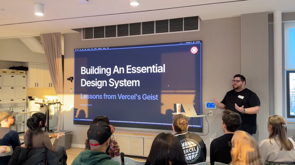 Alt text:   A speaker giving a presentation titled "Building An Essential Design System: Lessons from Vercel's Geist" to an audience in a modern conference room. The presenter stands to the right of a large screen displaying the presentation title, with a laptop and a drink on a small table beside him. The audience is seated facing the screen, attentively watching the talk. The room has light-colored walls, a window on the right, and storage cabinets on the left.