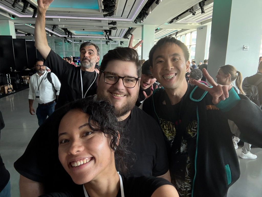 Alt text:  A group selfie of four smiling people indoors at a busy event or conference. In the foreground, a woman with curly hair smiles at the camera. Behind her, a man with glasses and a beard smiles, next to an Asian man flashing a peace sign with both hands. In the background, another man raises one arm enthusiastically while others walk and chat in the spacious, well-lit venue.