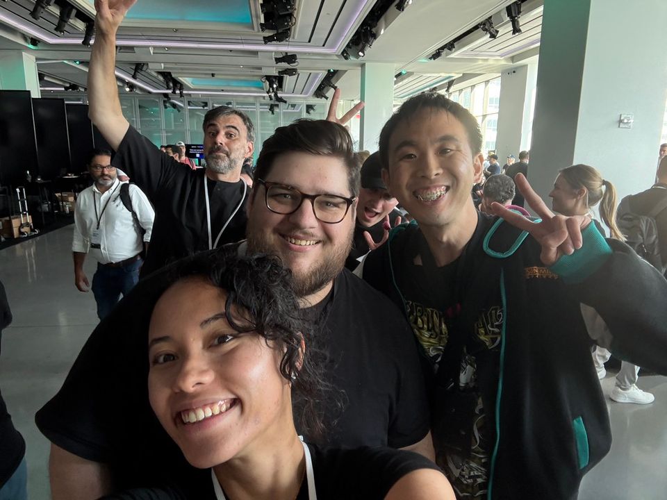 Alt text:  A group selfie of four smiling people indoors at a busy event or conference. In the foreground, a woman with curly hair smiles at the camera. Behind her, a man with glasses and a beard smiles, next to an Asian man flashing a peace sign with both hands. In the background, another man raises one arm enthusiastically while others walk and chat in the spacious, well-lit venue.