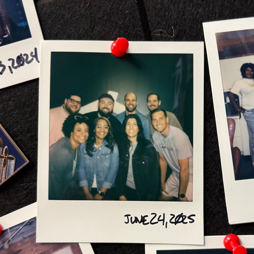 Alt text:  A pinned Polaroid photo showing a group of eight smiling people posing together against a dark background. The group consists of both men and women, dressed casually. The photo is dated "June 24, 2025" handwritten at the bottom. The Polaroid is pinned to a black fabric board with a red pushpin. Other pinned photos are partially visible around it.