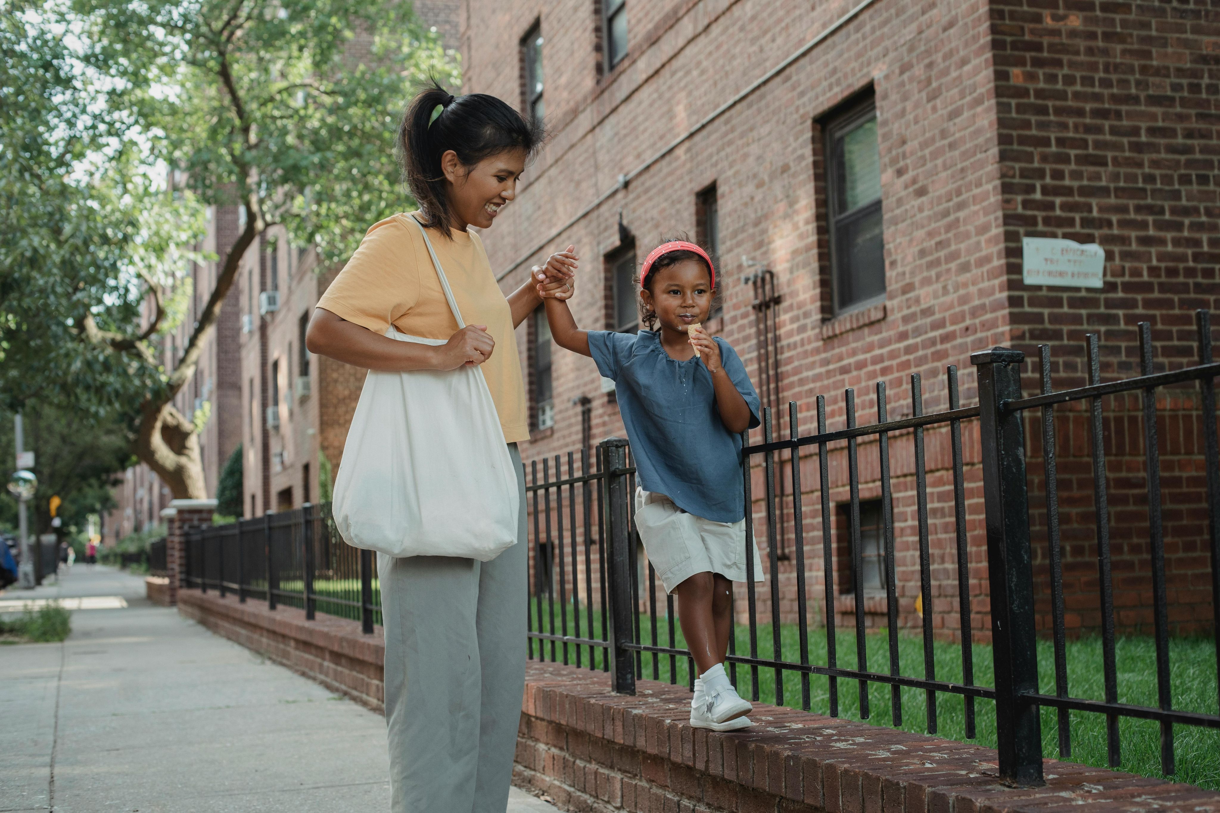 A woman holds a young child's hand as the child balances along a low brick wall on a city street.