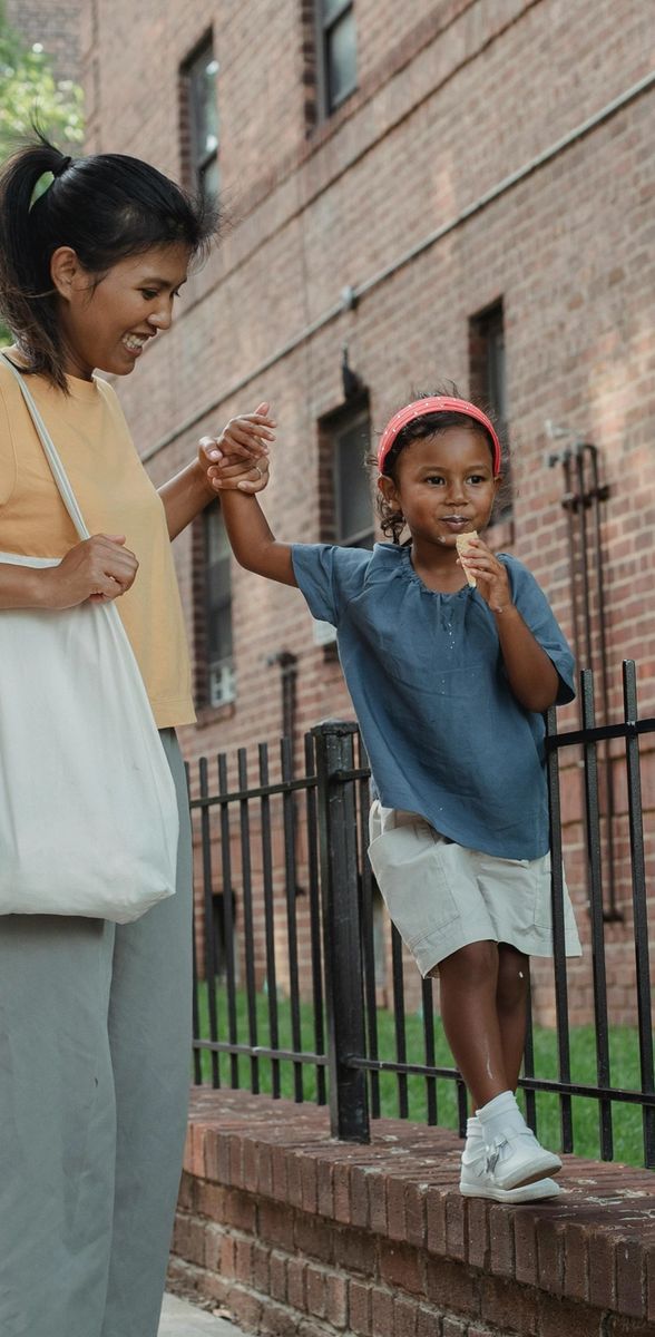 A woman holds a young child's hand as the child balances along a low brick wall on a city street.