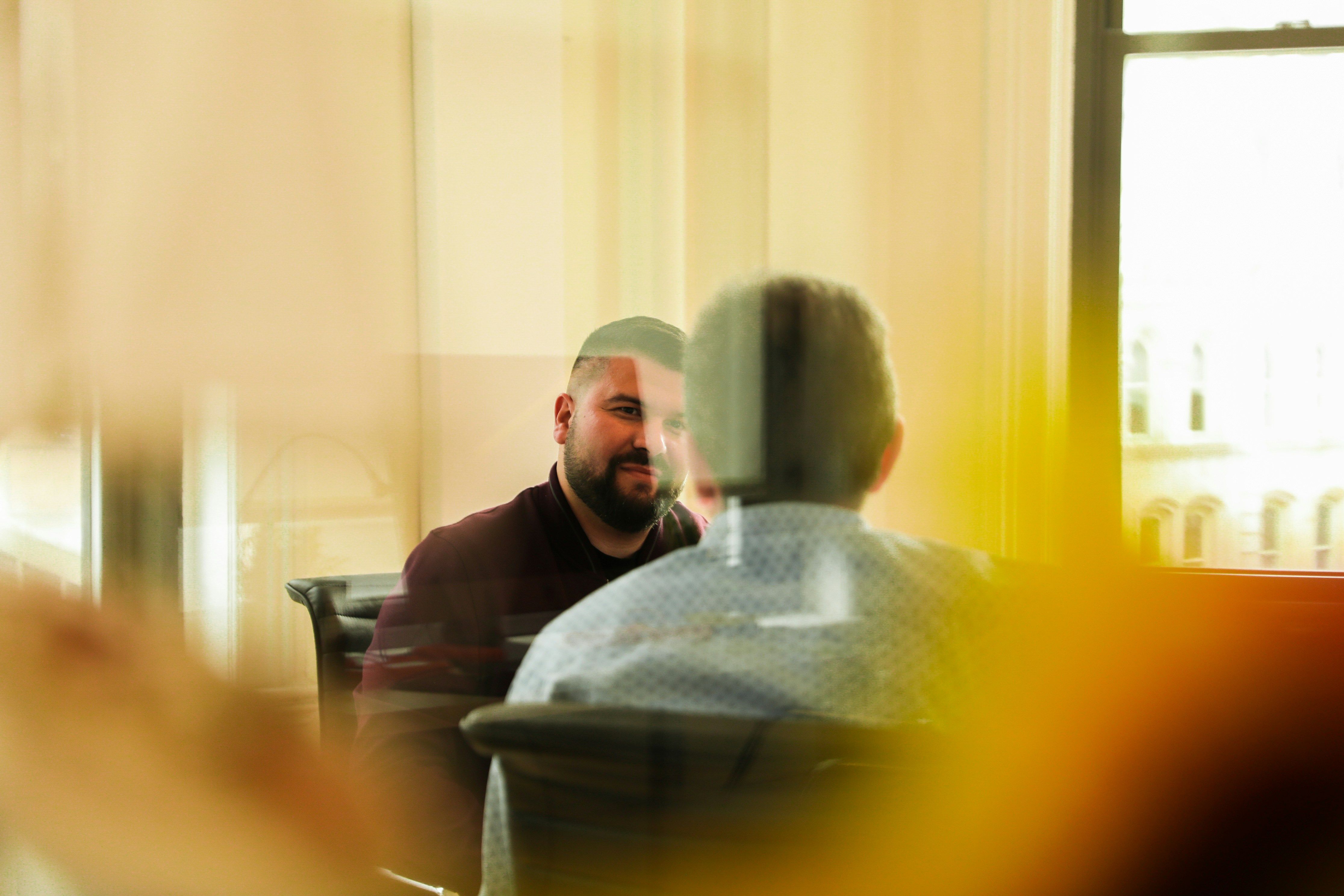 Two men sit facing each other in conversation, seen through a reflective window.