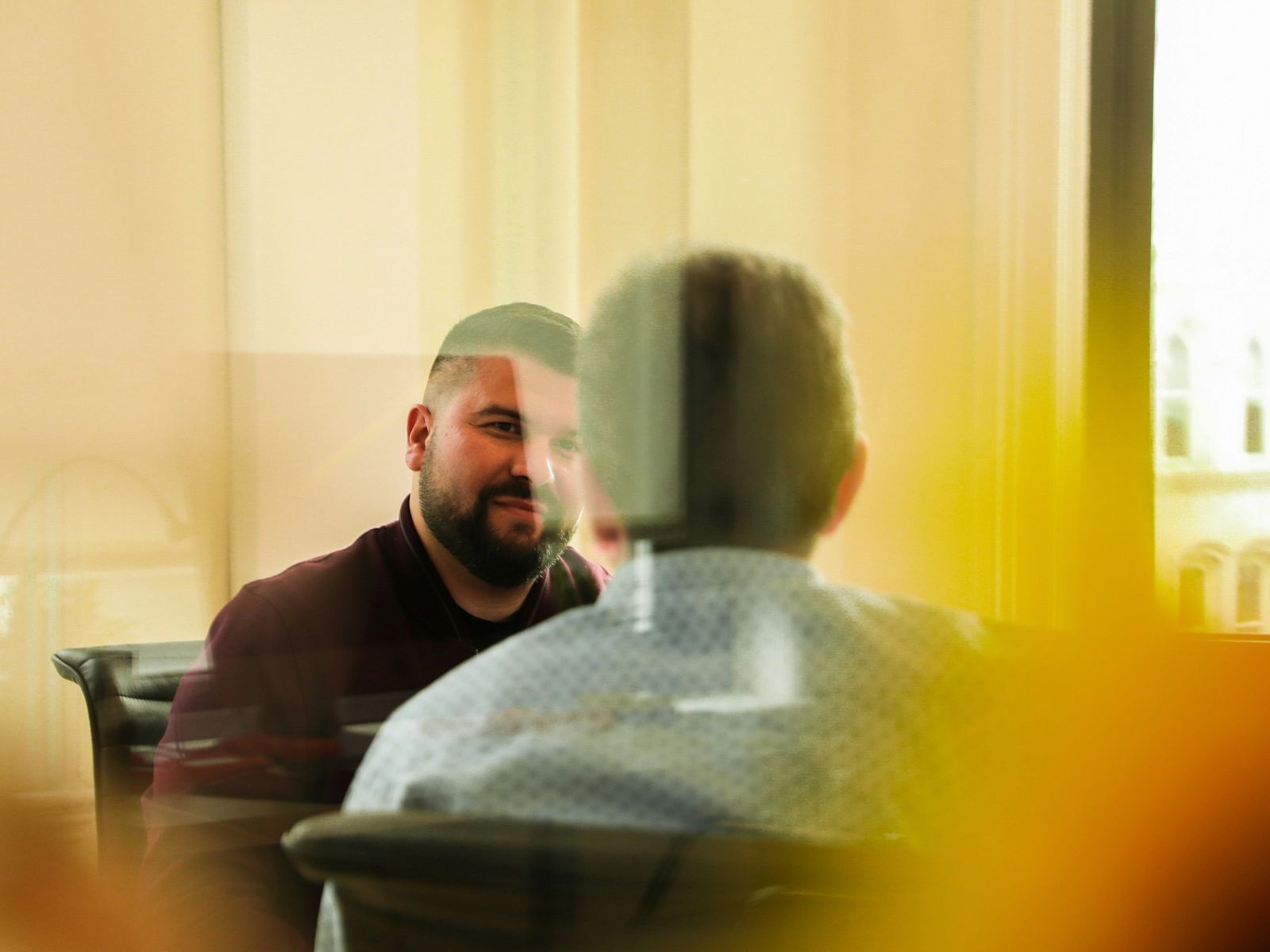 Two men sit facing each other in conversation, seen through a reflective window.