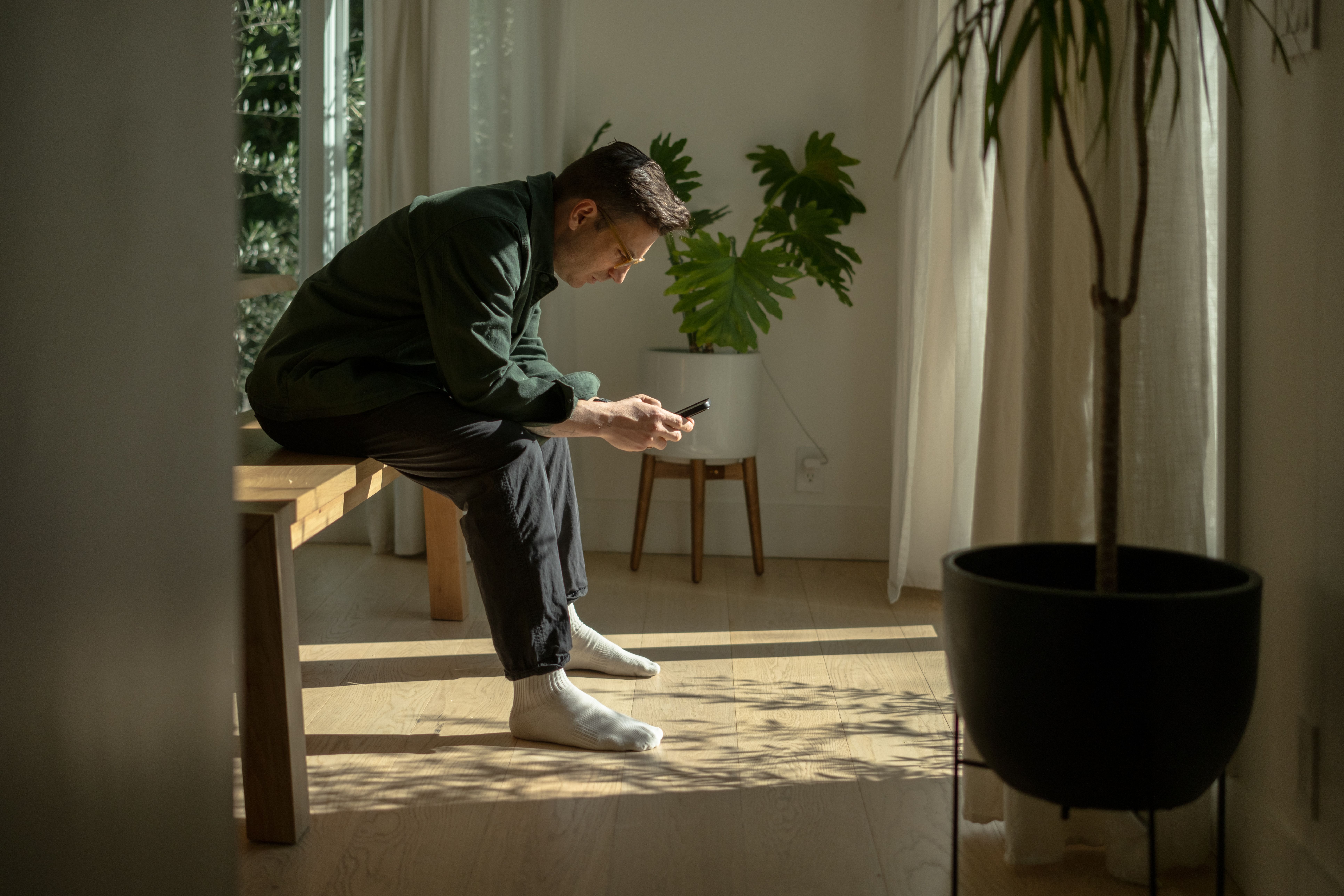 A man sits on the edge of a wooden bench in a sunlit room, looking down at his phone.