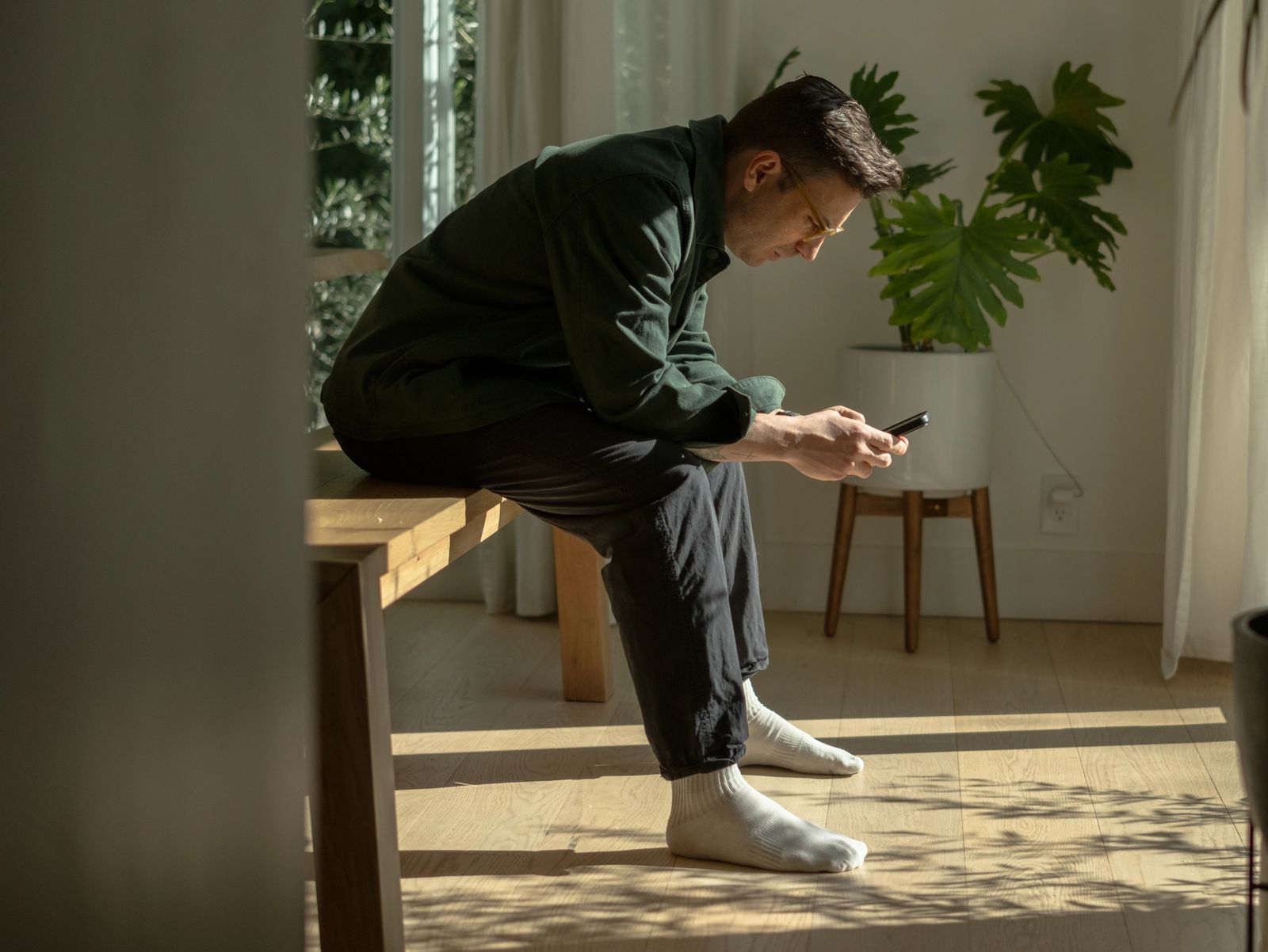 A man sits on the edge of a wooden bench in a sunlit room, looking down at his phone.