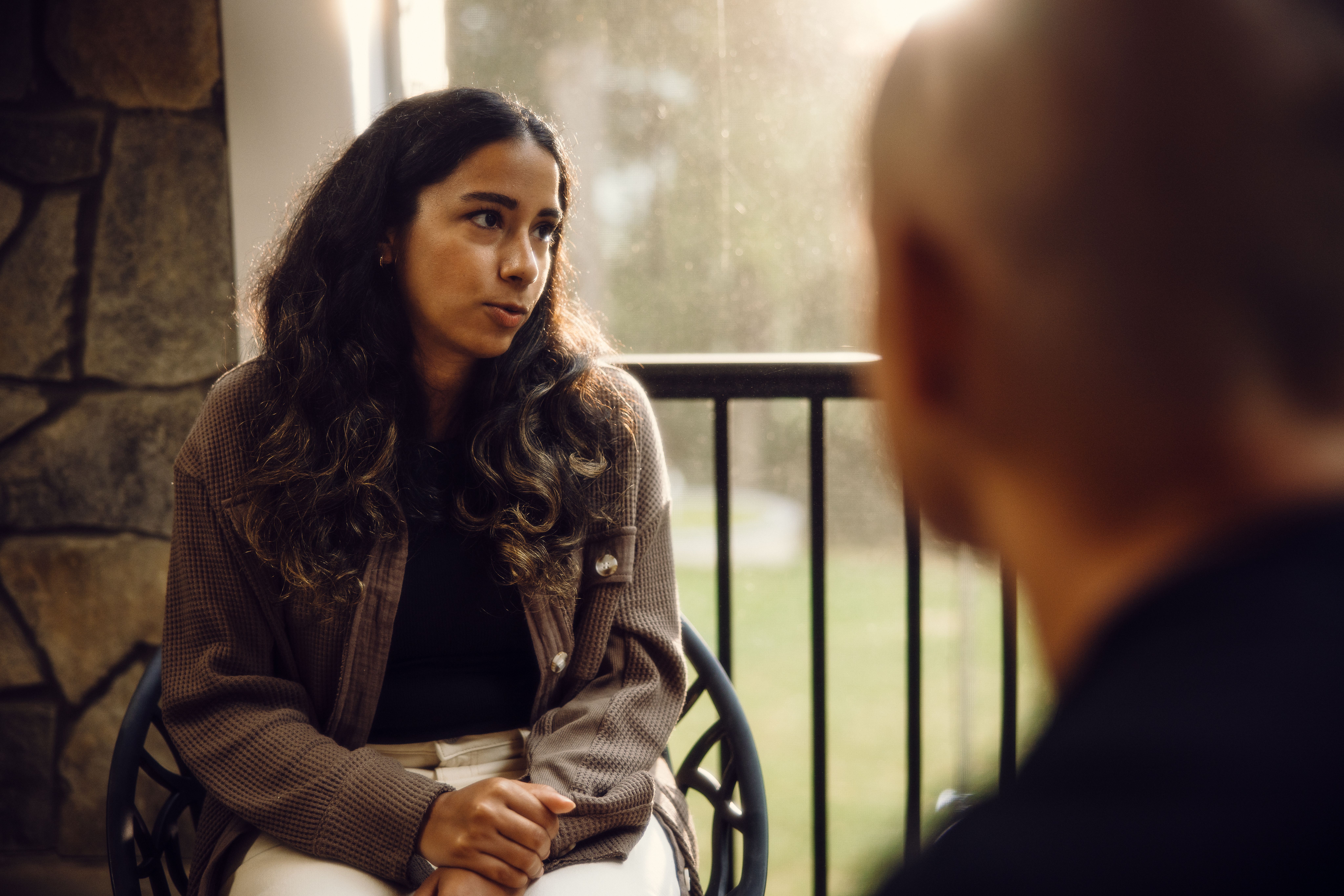 A young woman sits listening intently during a conversation with an older person on a porch.