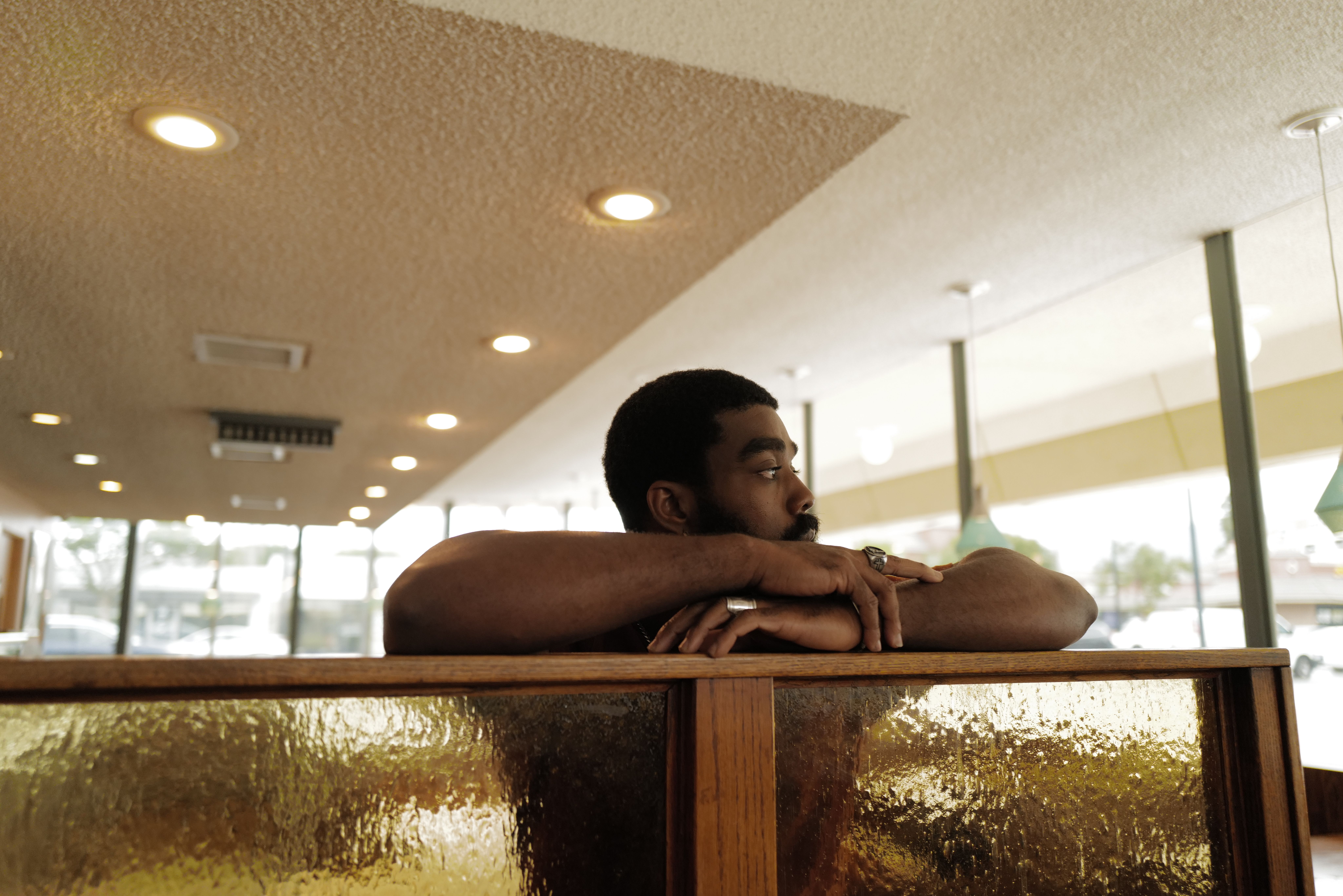 A man rests his folded arms on the back of a diner booth, looking out toward the window.