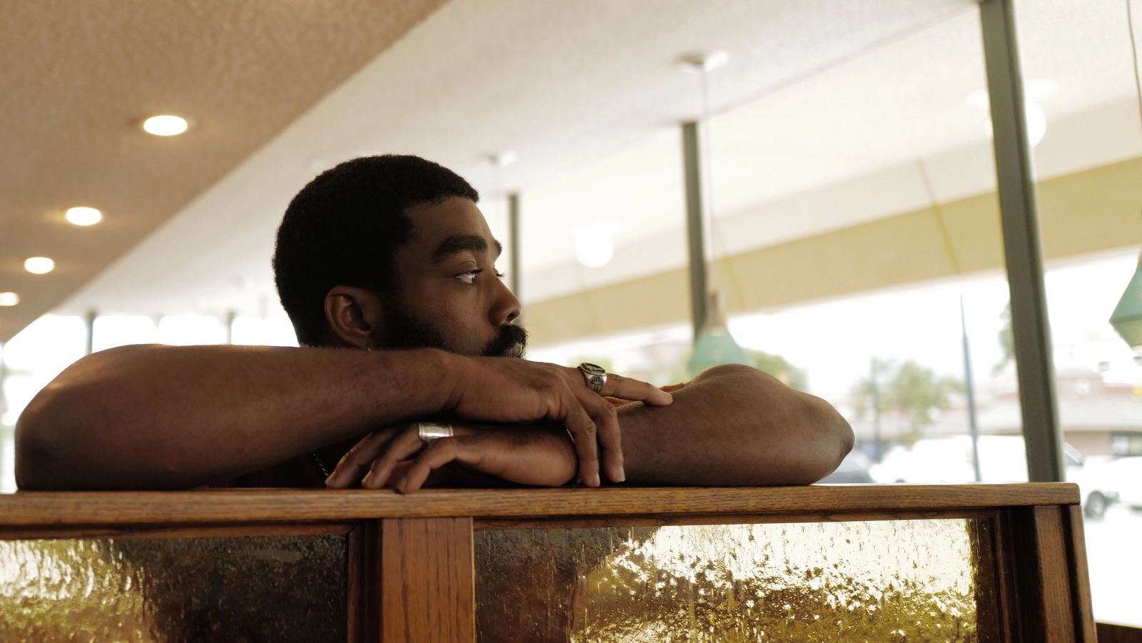 A man rests his folded arms on the back of a diner booth, looking out toward the window.