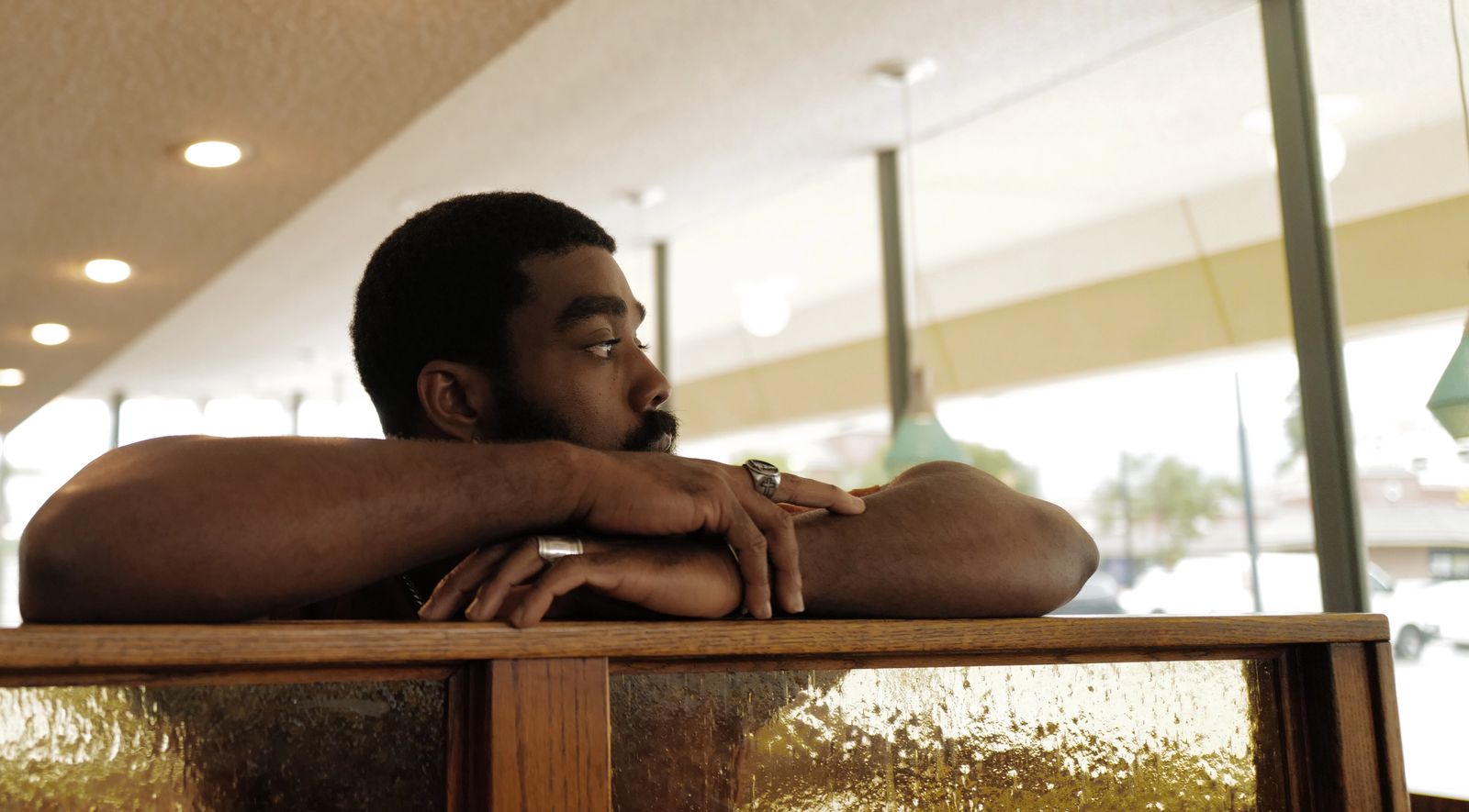 A man rests his folded arms on the back of a diner booth, looking out toward the window.