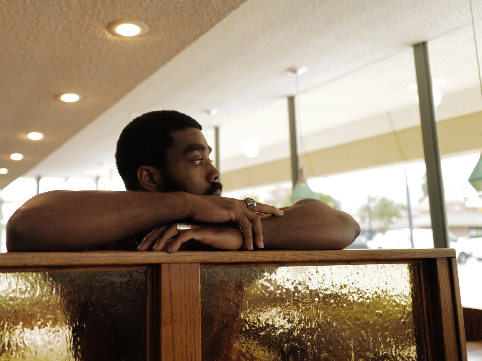 A man rests his folded arms on the back of a diner booth, looking out toward the window.