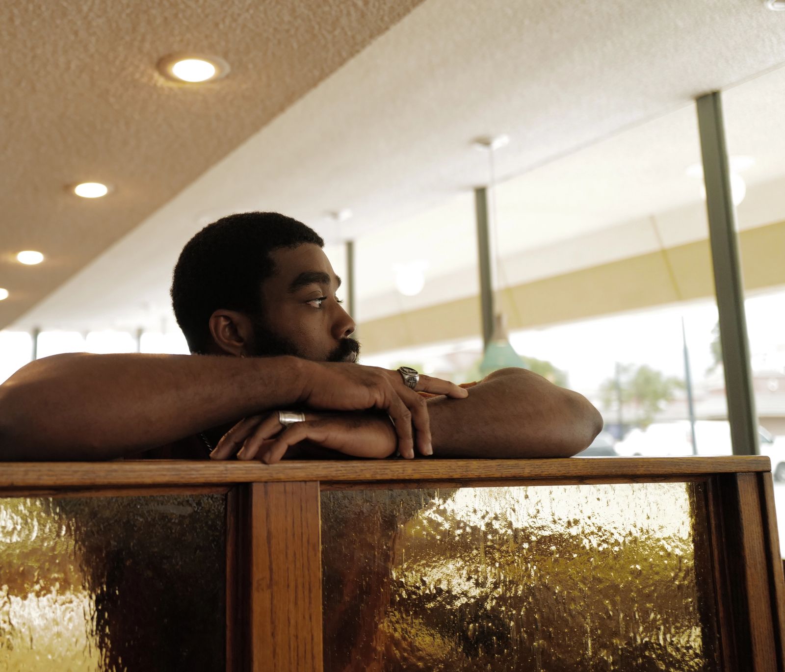 A man rests his folded arms on the back of a diner booth, looking out toward the window.