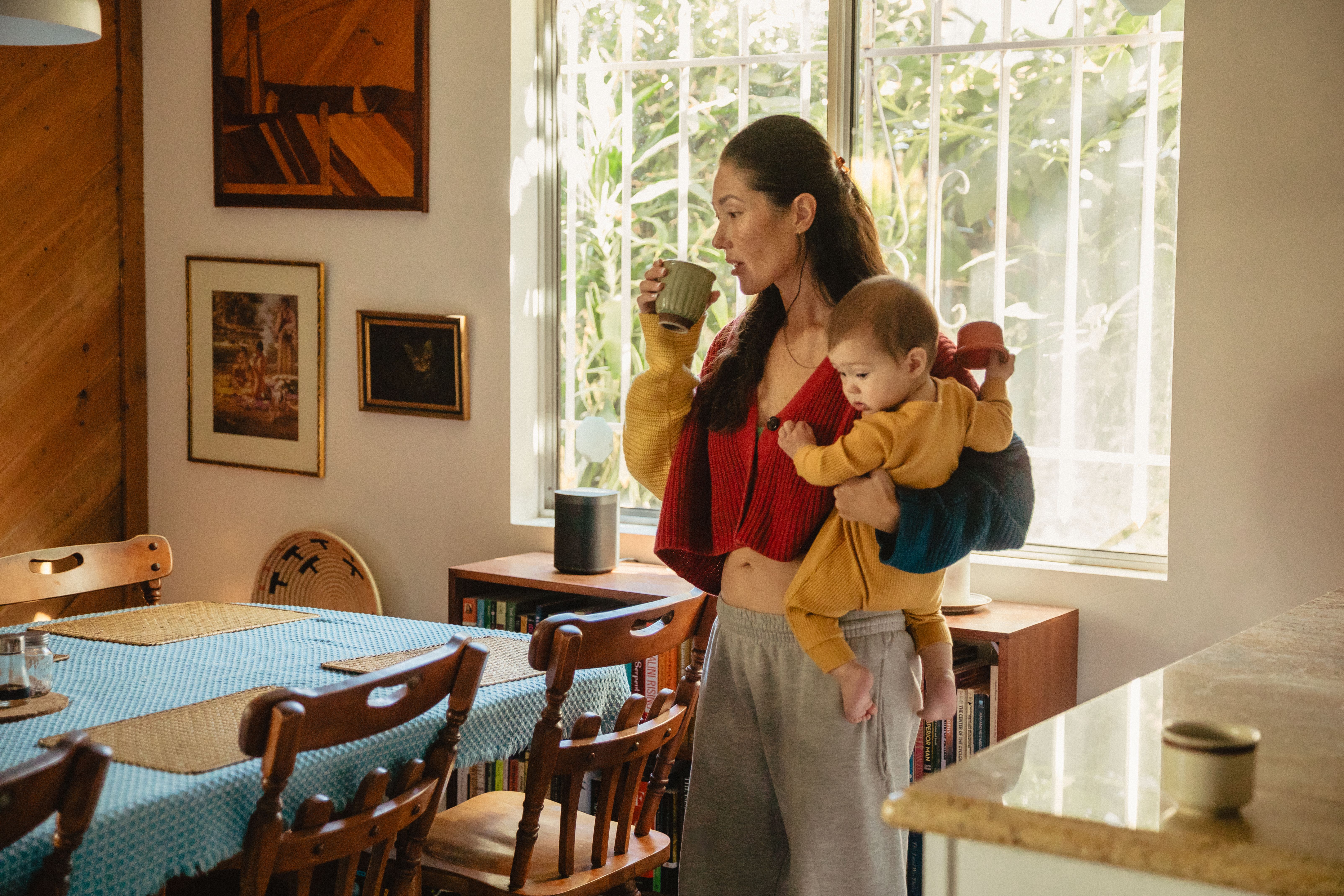 A woman drinks from a mug while carrying a baby on her hip in a sunlit dining room.