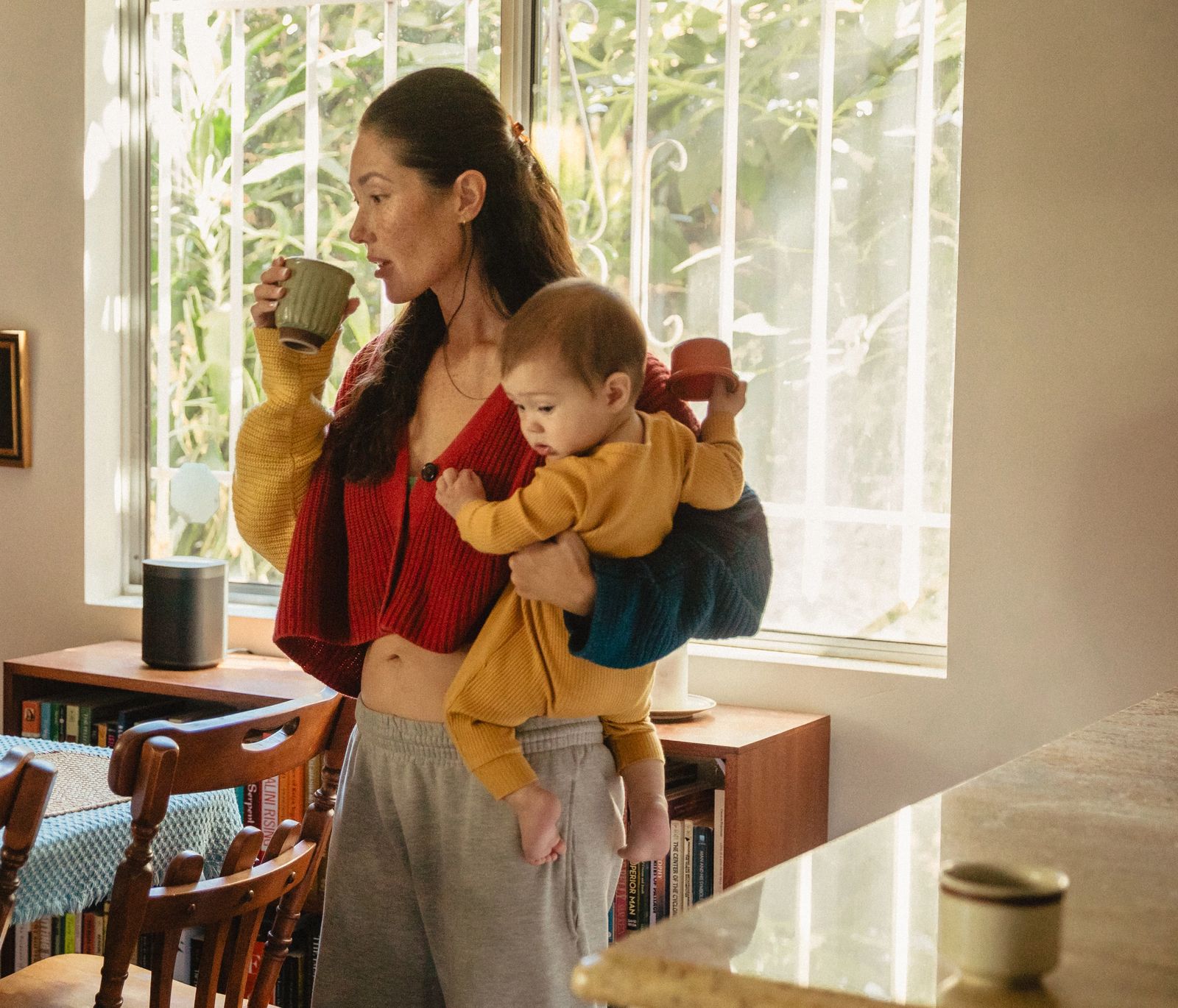 A woman drinks from a mug while carrying a baby on her hip in a sunlit dining room.