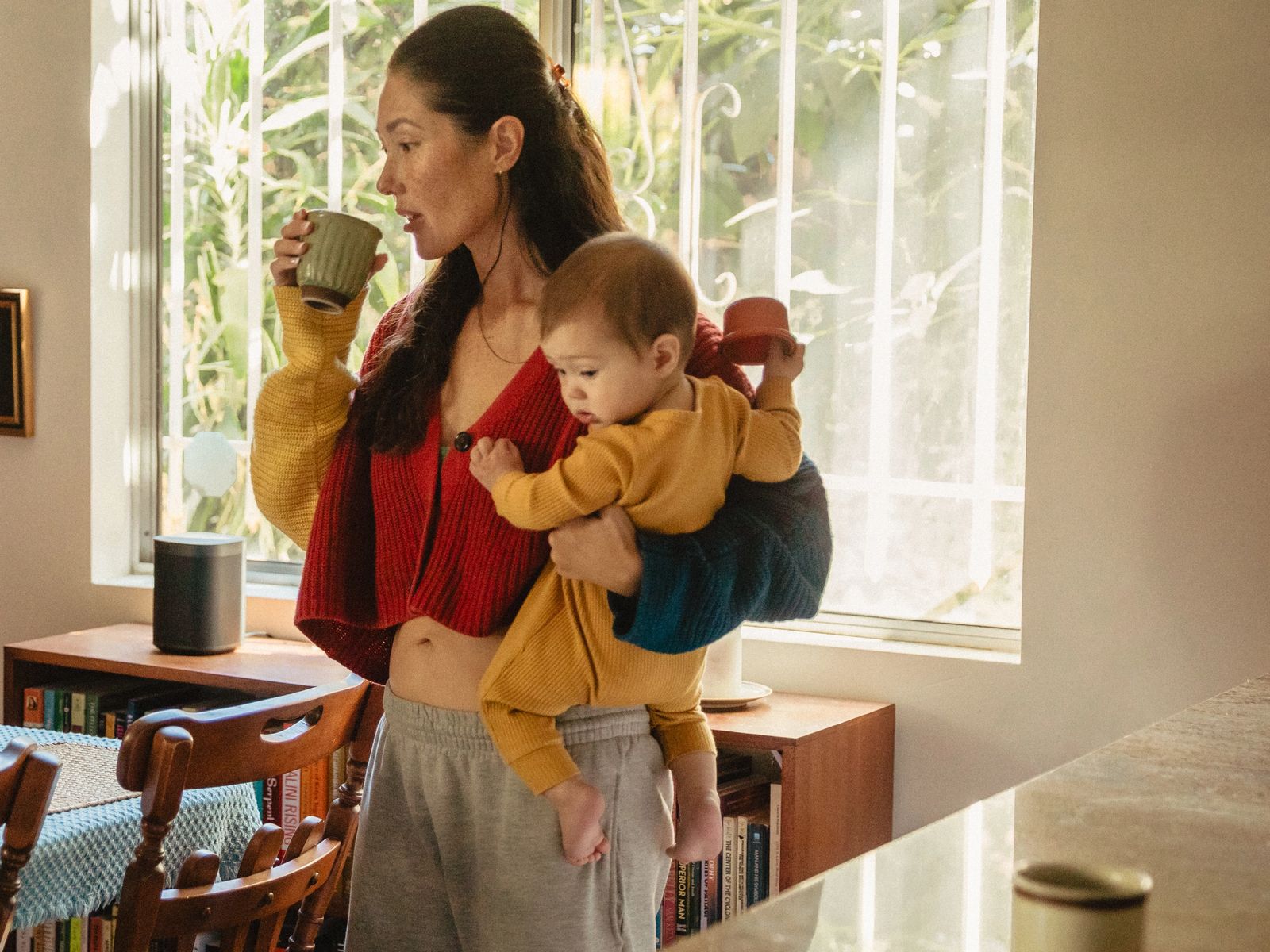 A woman drinks from a mug while carrying a baby on her hip in a sunlit dining room.