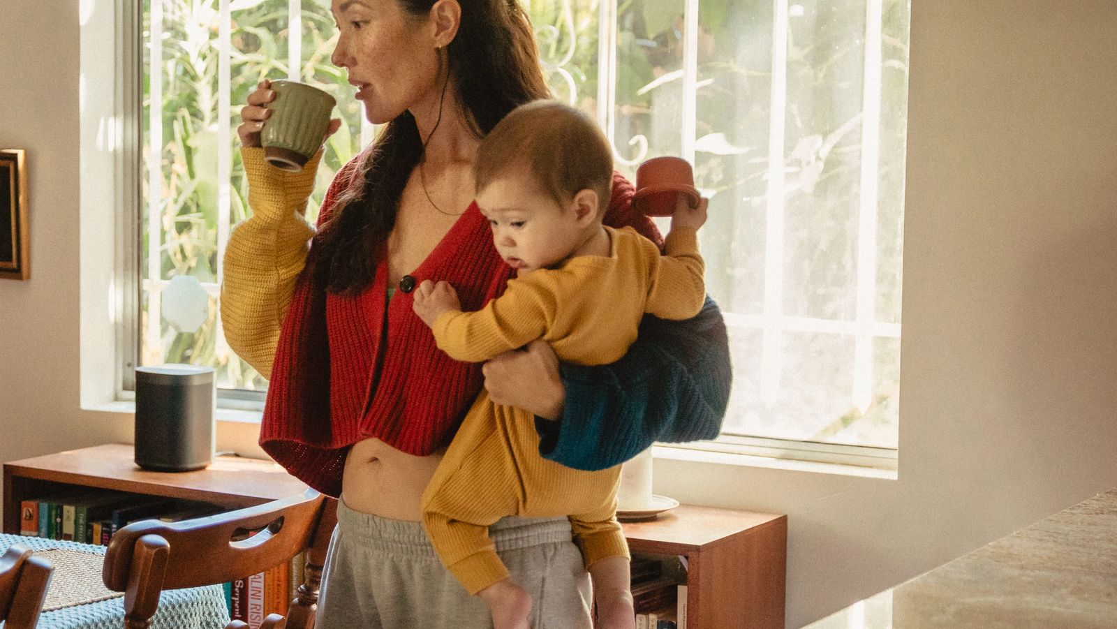 A woman drinks from a mug while carrying a baby on her hip in a sunlit dining room.