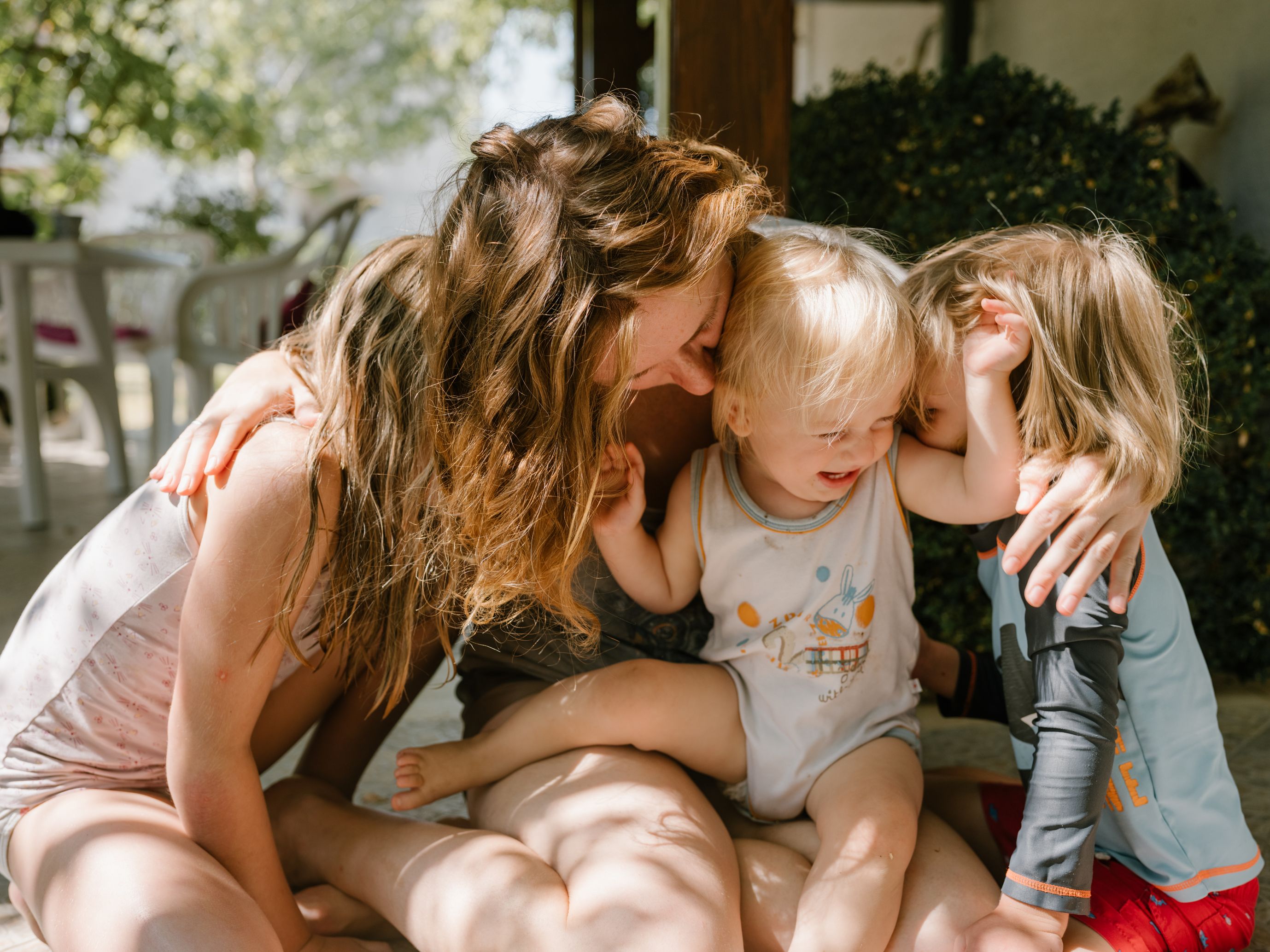 A woman sits on the ground with two young children draped over her, all of them laughing.
