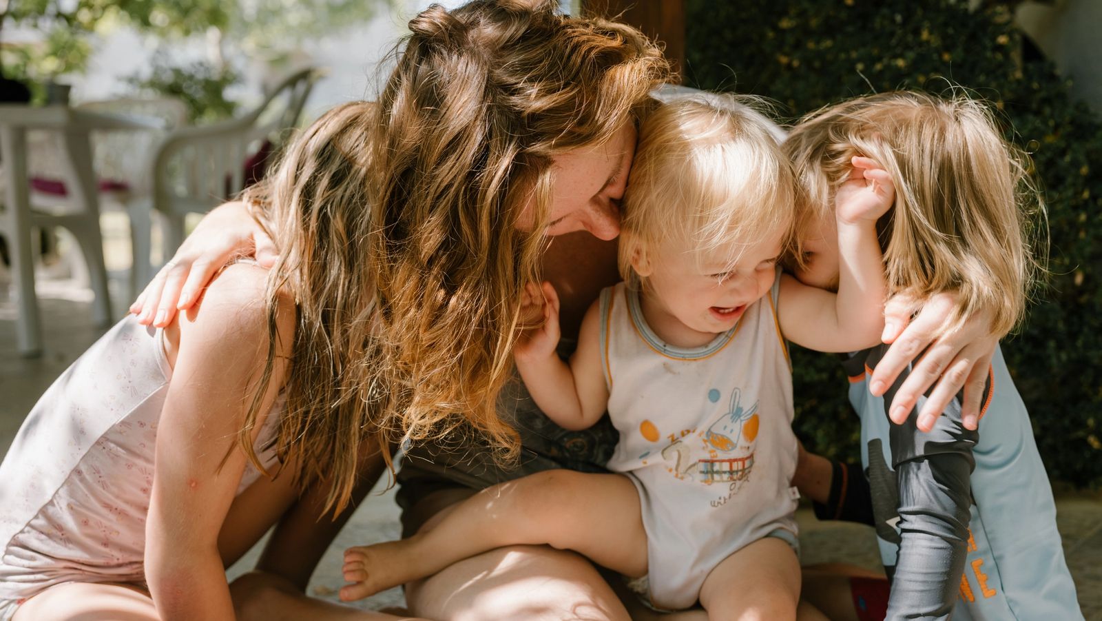 A woman sits on the ground with two young children draped over her, all of them laughing.