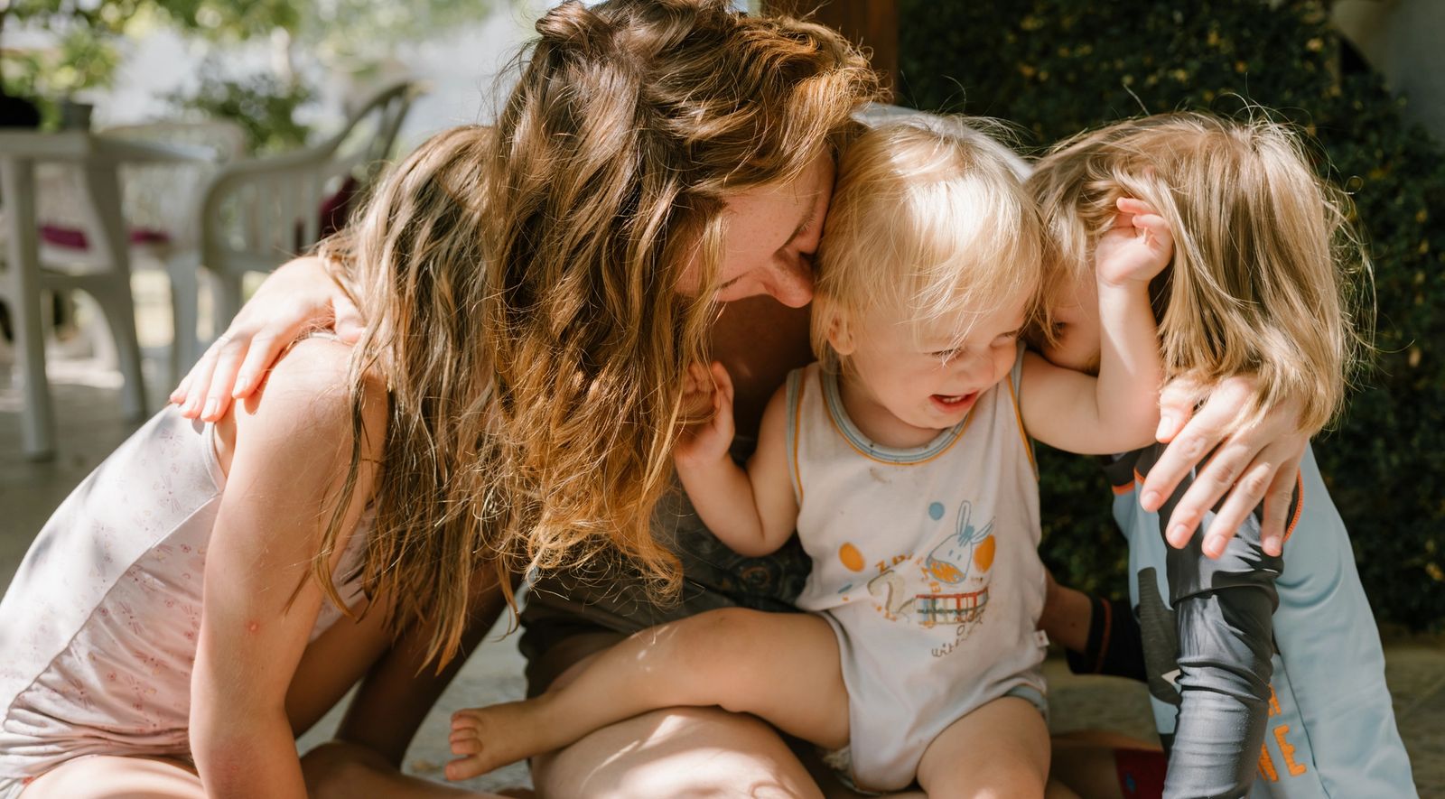 A woman sits on the ground with two young children draped over her, all of them laughing.